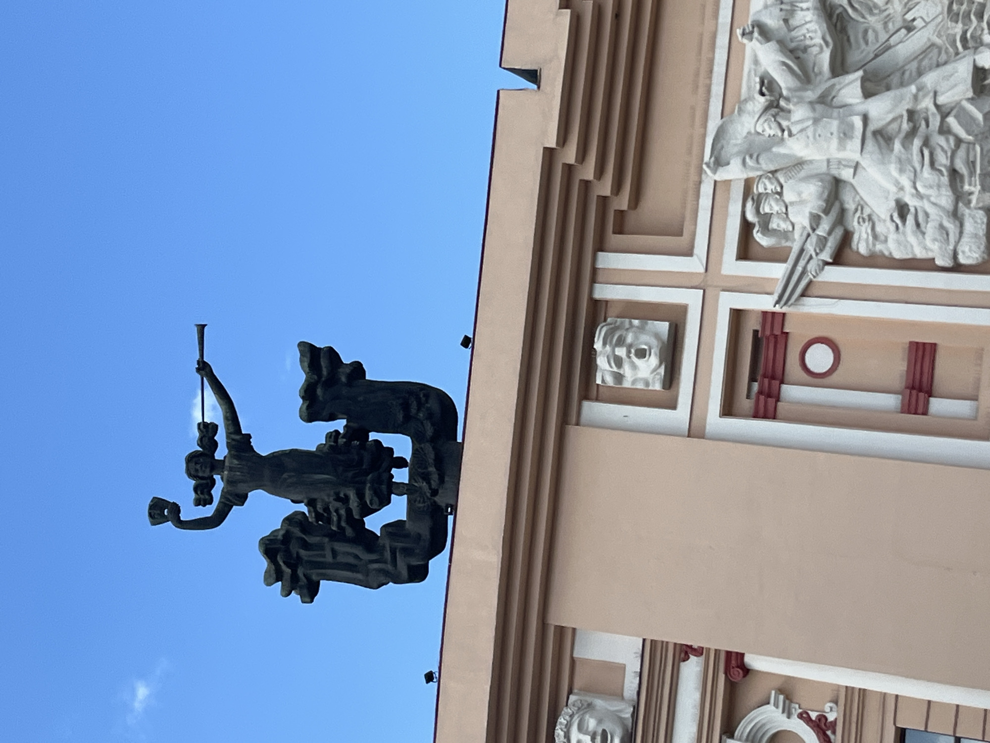 This is a vertical, eye-level, outdoor shot of a building with architectural details and a sculpture against a clear blue sky. The building, likely a monument or cultural center, features a beige facade with decorative elements. Prominently displayed on the building is a large, detailed white relief sculpture depicting several figures in what appears to be a dramatic or historical scene, possibly involving struggle or triumph. Below this, there are smaller relief sculptures of human faces set within square frames. Some sections of the building have red and white rectangular decorative elements.
To the left of the building, a dark-colored sculpture of a female figure is suspended in the air against the sky. She appears to be holding a horn to her lips, as if playing it, and has a flowing garment. The sculpture is dynamic and possibly represents a muse or a herald.
The scene is illuminated by natural light, suggesting it is daytime. The clear blue sky with a few wispy clouds indicates fair weather. The architectural style and the subject matter of the sculptures, particularly the relief depicting multiple figures in a narrative, are reminiscent of Soviet-era public art. While the location is identified as Dnipro, Ukraine, there are no visible signs or text in the image that can provide specific clues about the name of the building or the sculptures. The overall impression is one of public art and historical commemoration.