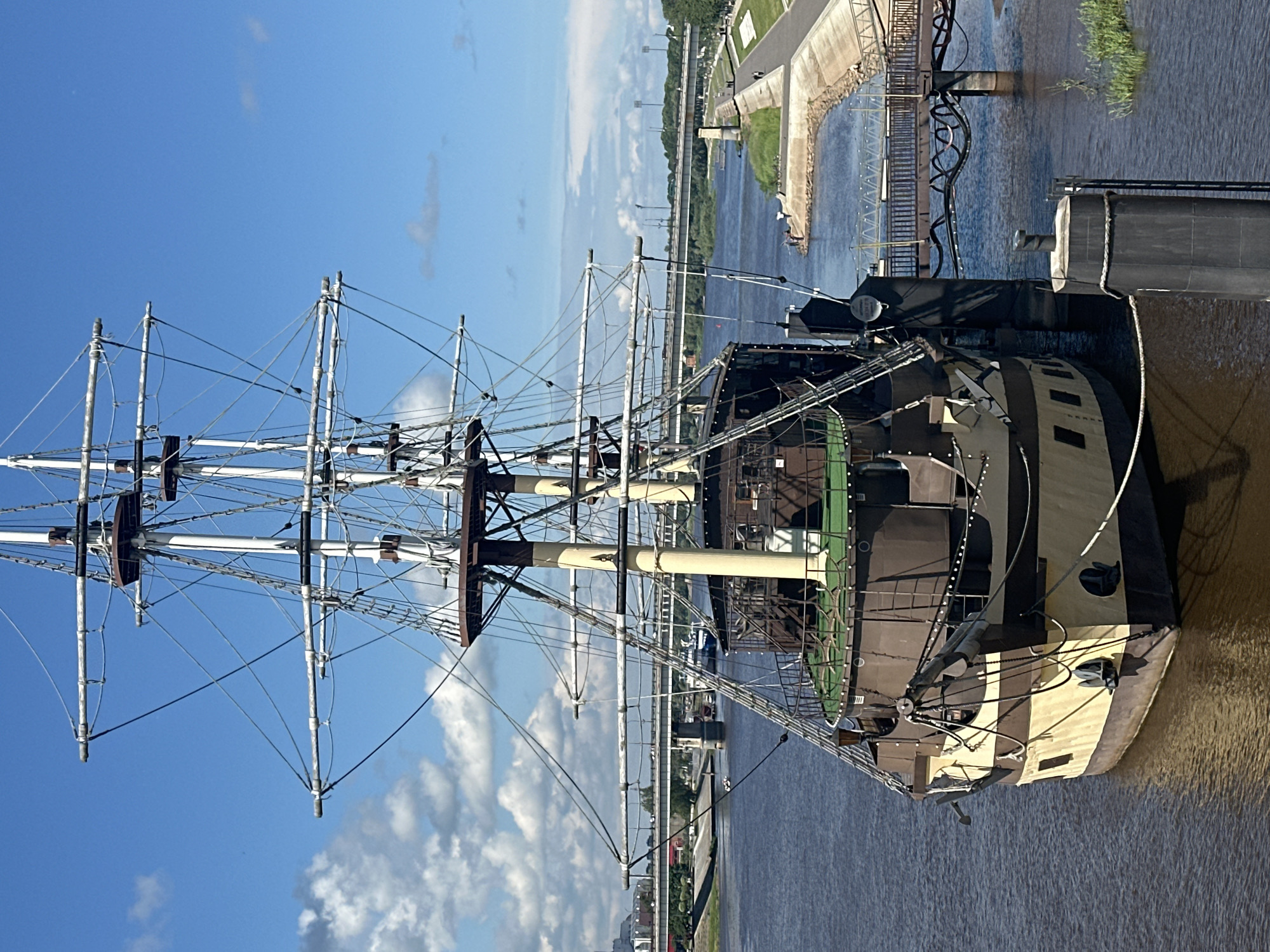 The image captures a large, multi-masted sailing ship docked on a wide, calm river. The ship, painted in brown and cream colors, has a prominent bow adorned with anchors and ropes. Its tall masts are intricately rigged with numerous lines, suggesting a historical replica or a vessel designed for show. The riverbank is lined with a paved walkway, a concrete embankment, and grassy areas. In the background, a long, multi-span bridge crosses the river, with cityscape visible on the far bank. The sky is bright blue with scattered white clouds, indicating a clear, sunny day. The overall scene depicts a picturesque waterfront setting, likely a tourist attraction or a port in Veliky Novgorod, Russia, given the distinctive bridges and urban architecture that are characteristic of the city. There are a few people visible on the riverbank walkway in the distance, and some individuals fishing near the embankment. The ship appears to be a static display, accessible via a walkway that extends from the shore.