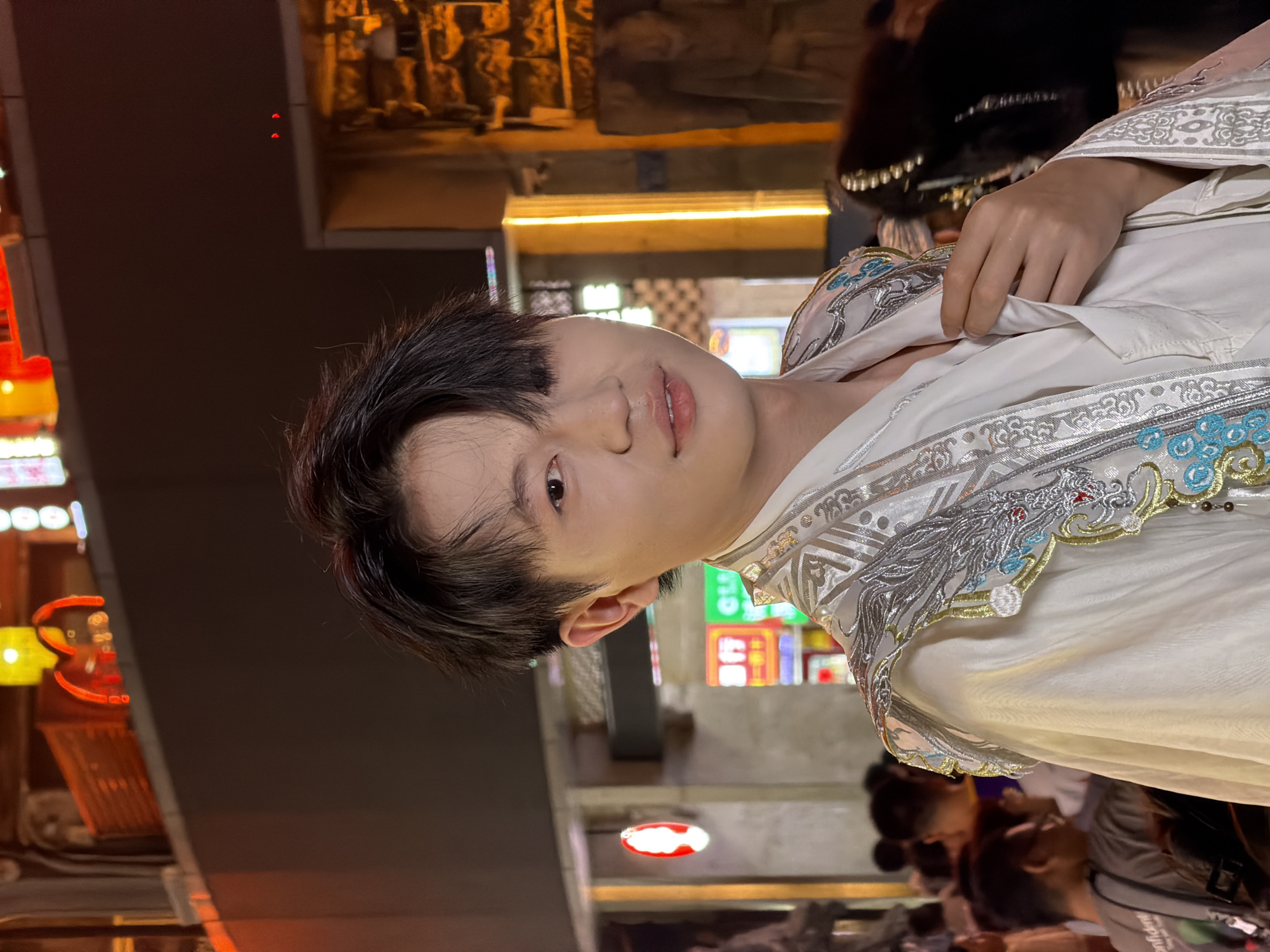 A young East Asian male with dark, slightly messy hair is positioned in the center of the frame, looking towards the camera. He is wearing a white traditional-style garment with ornate silver and blue embroidery featuring dragon motifs on the shoulders and collar. His right hand is slightly raised, touching the front of his garment.
The background suggests an urban setting at night, likely in Yuzhong District, China, characterized by bright neon signs, illuminated buildings, and a crowd of people. The lighting is dim, with yellow and orange hues from the ambient lights casting a warm glow. Several signs with Chinese characters are visible in the background, one in particular appearing to be a red circular logo with a silhouette of a face. The overall atmosphere is bustling and vibrant.