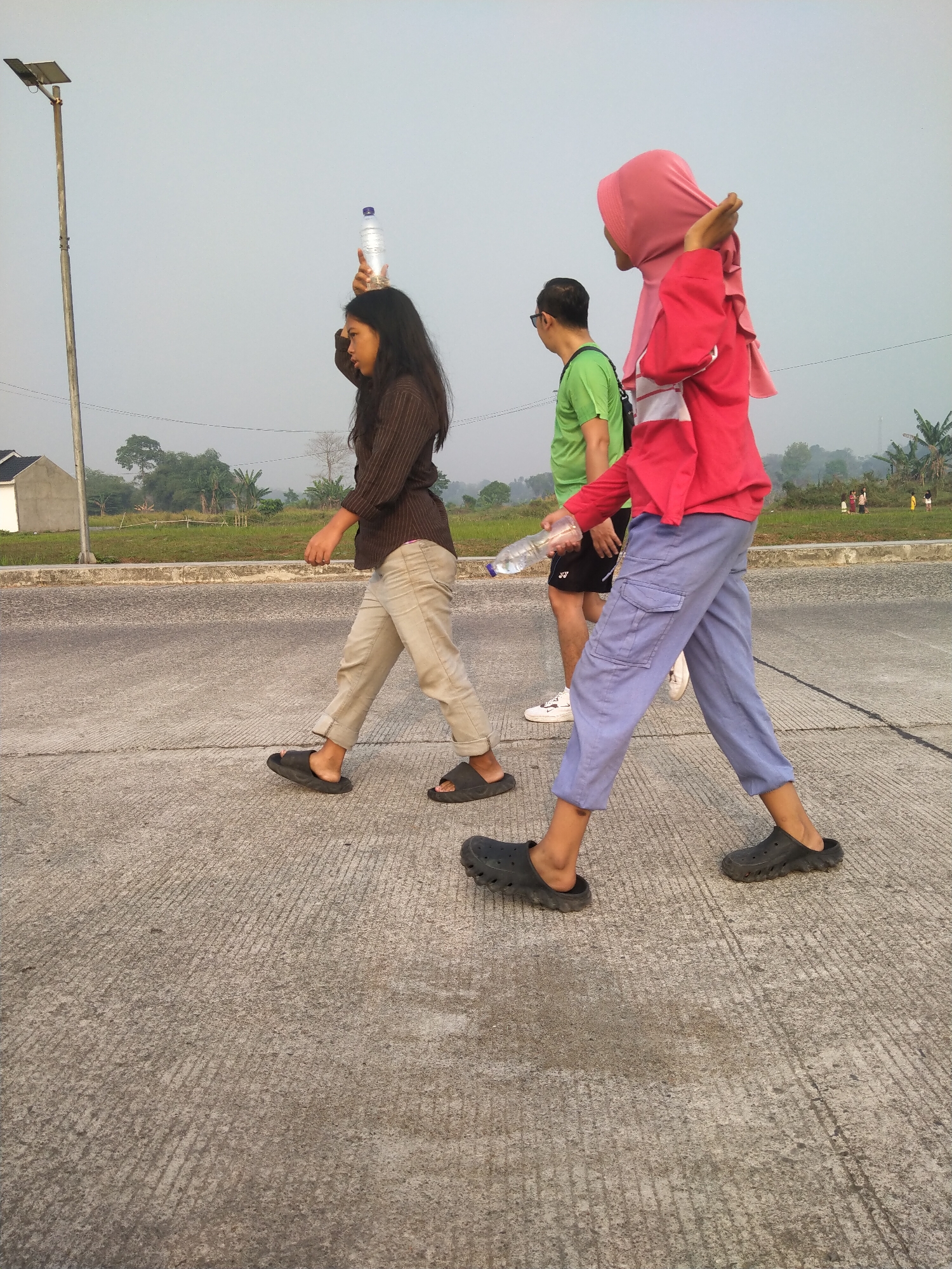 In Cisoka, Indonesia, three individuals are walking on a paved surface under a hazy sky. The person in the foreground, on the right, wears a bright pink hijab and a matching long-sleeved top with white accents, paired with loose-fitting light blue pants and black clogs. They are holding a clear plastic water bottle in their right hand. To their left, a person with dark hair, wearing a brown striped long-sleeved shirt, light-colored pants, and black slides, walks with a clear plastic water bottle balanced on their head. Further to the left, a person in a green t-shirt and black shorts, wearing white sneakers, walks with a black bag slung over their shoulder. In the background, there is a solar-powered street lamp, a small white building, and a field with scattered trees. The ground is a light gray concrete with visible expansion joints and some staining.