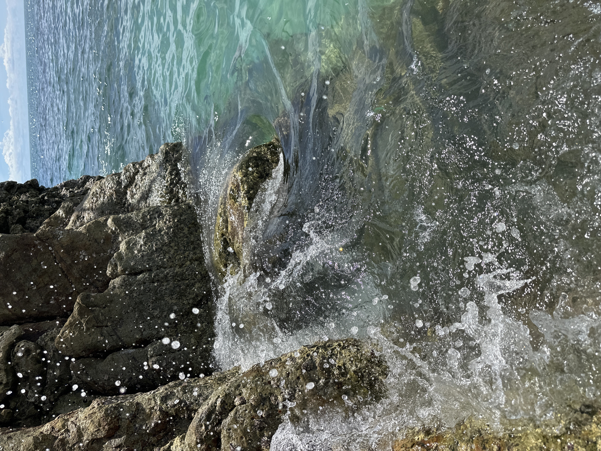 This is a close-up, eye-level shot of waves crashing against rocks at the edge of the ocean. The water is clear and turquoise, with sunlight glinting off the surface. The rocks are rough and brown, covered in small barnacles. Droplets of water are suspended in the air, creating a dynamic and refreshing effect. The background shows the vast expanse of the sea under a bright blue sky with scattered white clouds. The scene is set in ตำบลเกาะพะงัน, Thailand, suggesting a tropical coastal environment.