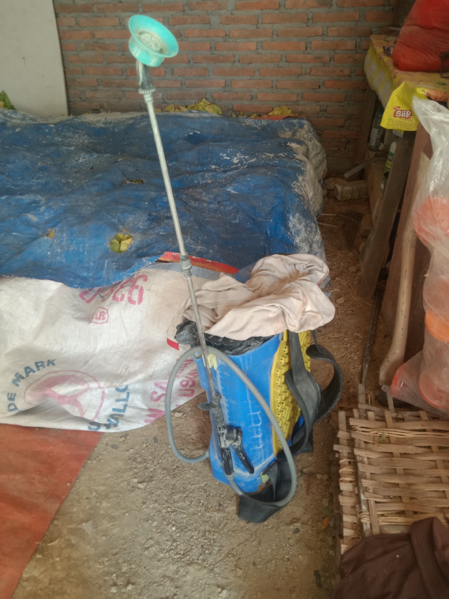 In Pacing, Indonesia, a backpack sprayer with a blue tank and yellow grate sits on the ground. A white bag with "DE MARK" and other lettering is partially visible. A blue tarp is draped over something behind the sprayer. A brick wall forms the background, and a woven mat is to the right. Various items, including what appears to be a red object in a plastic bag, are stacked to the right of the sprayer. The ground is covered with dirt and gravel.