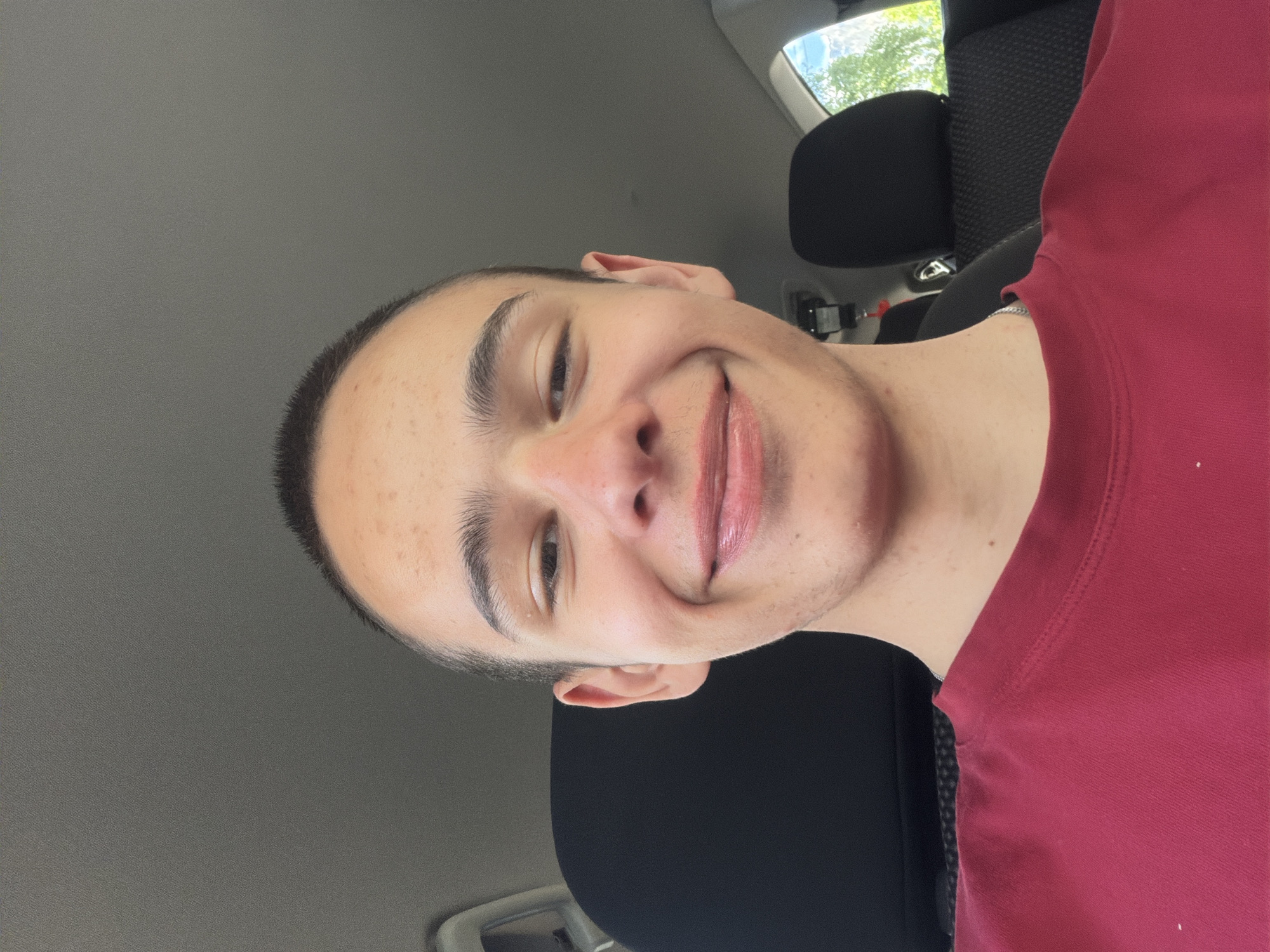 A close-up selfie shows a young person with a buzz cut, wearing a red t-shirt, smiling. They are seated in a car, with dark headrests visible in the background. The lighting suggests an outdoor setting, possibly daytime. The car interior appears to be gray. The person's skin has some light freckling on the forehead and cheeks. They are wearing a silver chain necklace.