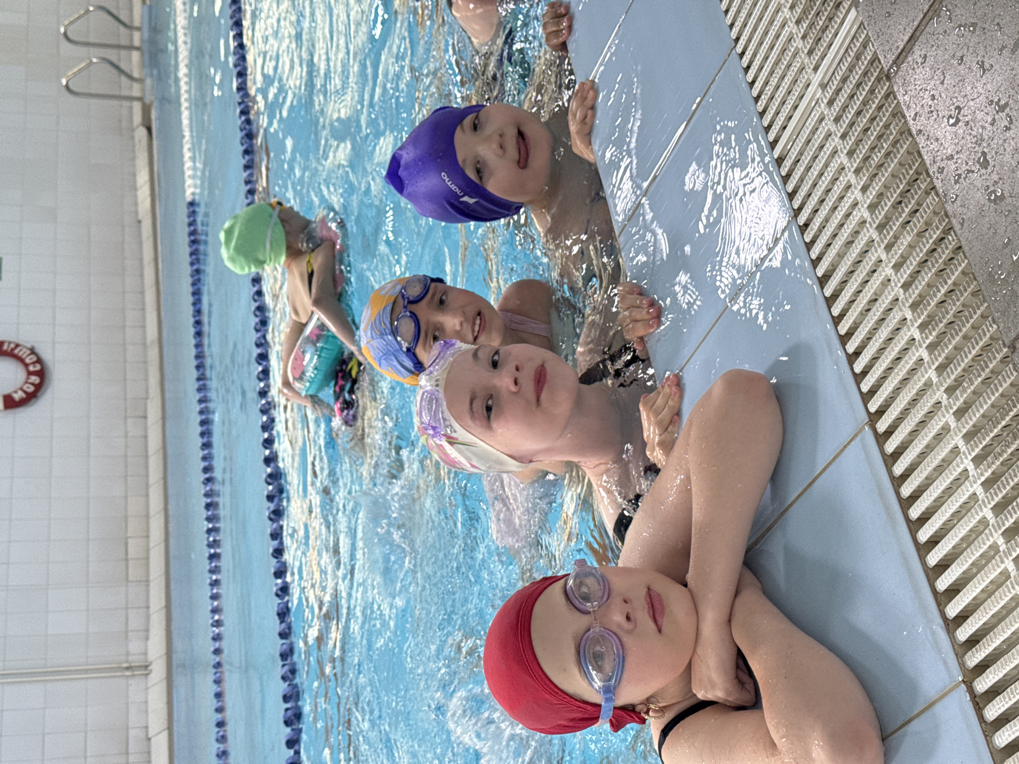 A group of young children are participating in a swimming lesson in an indoor pool. The children are wearing swim caps and goggles, and some are holding onto the side of the pool. The pool is a bright blue, with lane lines visible. The walls of the pool are tiled in white and light blue. There is a red life preserver ring visible on the left side of the image. The children appear to be enjoying themselves, with some smiling and looking at the camera. The scene suggests a fun and educational activity for the children.