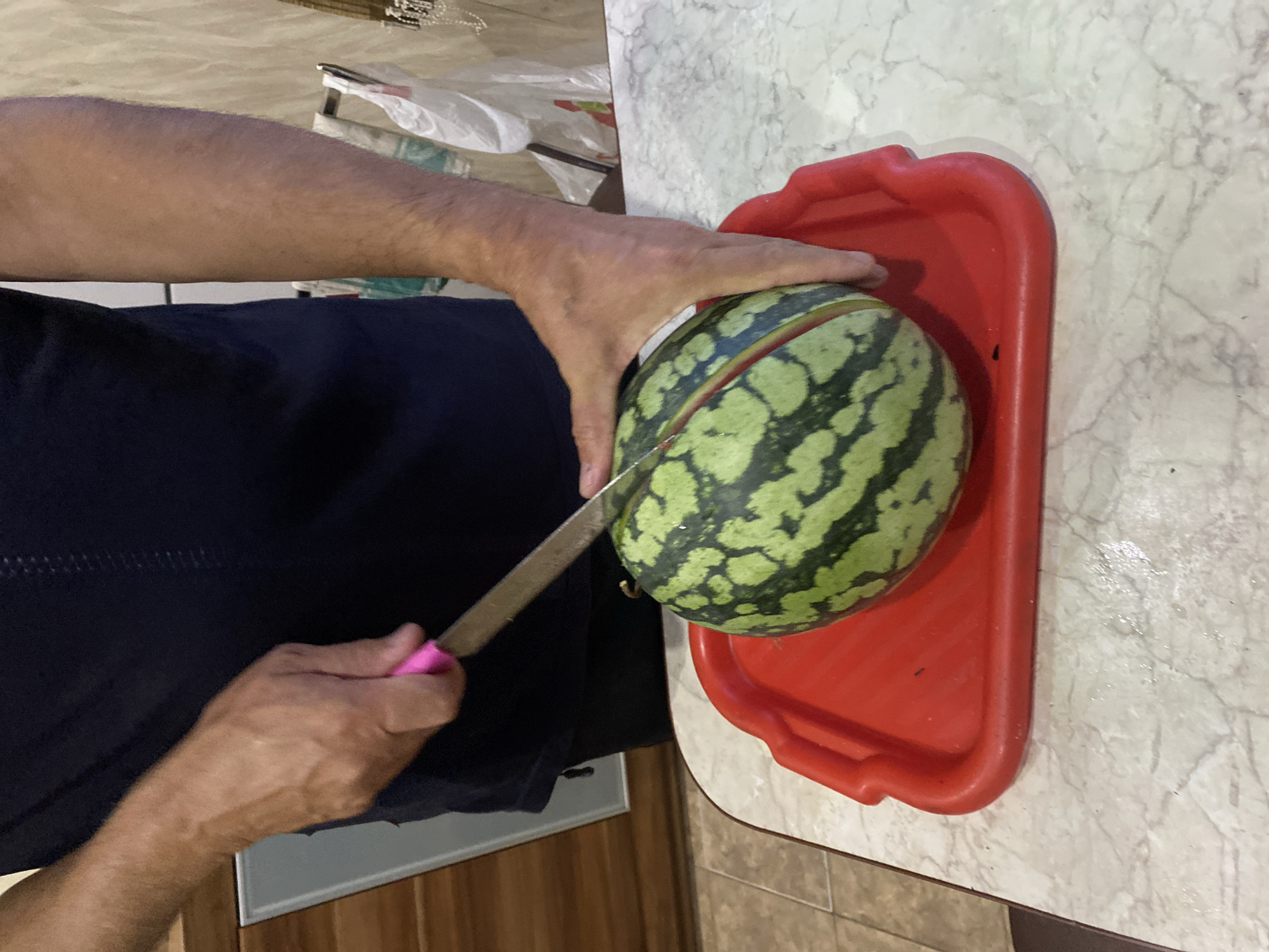 A person, whose gender is not discernible, is in the process of cutting a small, round watermelon with a knife that has a pink handle. The watermelon is placed on a red plastic tray, which is itself on a white marble countertop. The background is out of focus but appears to be a kitchen setting, with cabinets and some plastic bags visible. The person is wearing a dark blue t-shirt. The scene is indoors.