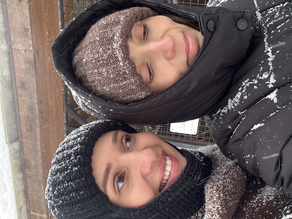 Two women, likely friends, are posing for a selfie outside in the snow. The woman at the bottom of the frame smiles widely, wearing a black knit hat and scarf. Her teeth are visible. The other woman wears a light brown knit hat and is partially obscured by the hood of a black winter coat. Both women have snow on their hats and coats. The background is a blurred mix of snowy wood and metal.
