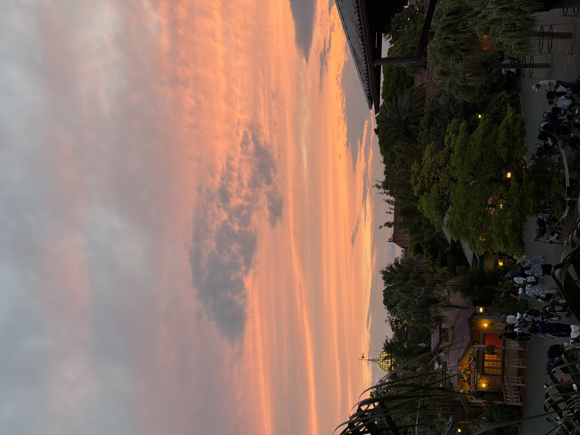The image captures a sunset scene in Tokyo Disneyland, Urayasu, Japan. The sky is filled with vibrant orange and pink hues, with scattered grey clouds. In the foreground, lush green foliage partially obscures the view. In the mid-ground, several buildings with rustic architecture are visible, including a prominent structure with a red roof and illuminated windows. A dome-shaped building with a yellow flag on top is also noticeable. A group of people, some with strollers, are walking around a paved area in front of the buildings. The overall atmosphere suggests a theme park setting during dusk.