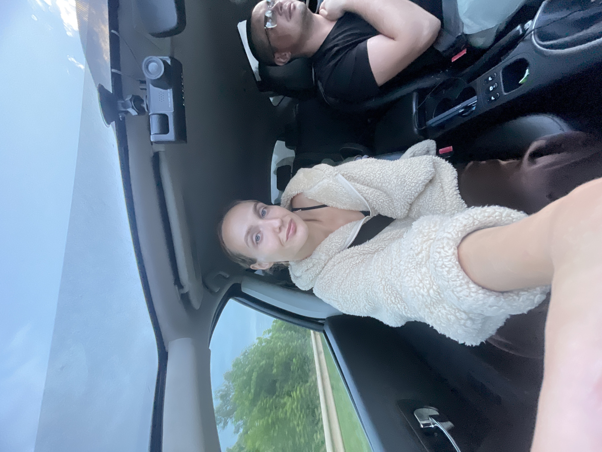 The image shows two people in the front seats of a car, likely traveling on a road in Pirogovo, Russia. The woman in the foreground is taking a selfie, wearing a fluffy cream-colored jacket over a black top. She has blonde hair tied back and is looking at the camera with a slight smile. To her right, in the passenger seat, a man wearing glasses and a black t-shirt is visible. He is looking forward, seemingly driving or in the passenger seat. The car's interior is visible, including the dashboard and a sunroof that lets in the daylight. Outside the car, lush green trees line the road, suggesting a rural or suburban setting. A dashboard camera is mounted near the sunroof.