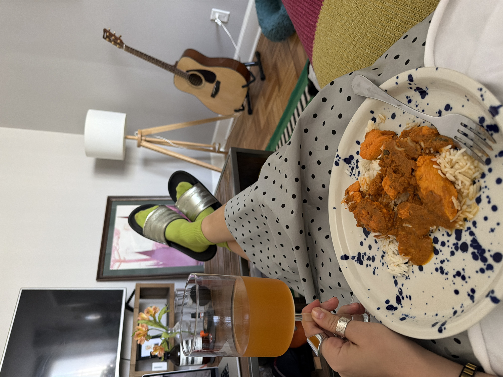 The image shows an indoor scene featuring a person seated on a colorful cushion. The person is wearing a gray polka-dot skirt, chartreuse socks, and silver sandals. They are holding a plate of food, which appears to be Indian cuisine consisting of rice and a curried dish, along with a fork. In the other hand, they are holding a glass of orange juice. Their fingers are adorned with multiple rings.
In the background, there is a guitar resting on a stand, a floor lamp, a framed picture, and a television set. A shelf with flowers is visible near the television.
Given the casual, home-like environment and the presence of personal items, the scene likely depicts a moment of relaxation at someone's residence. While specific markers indicating location in Los Angeles are absent, the setting could be in any residential area within the city.
