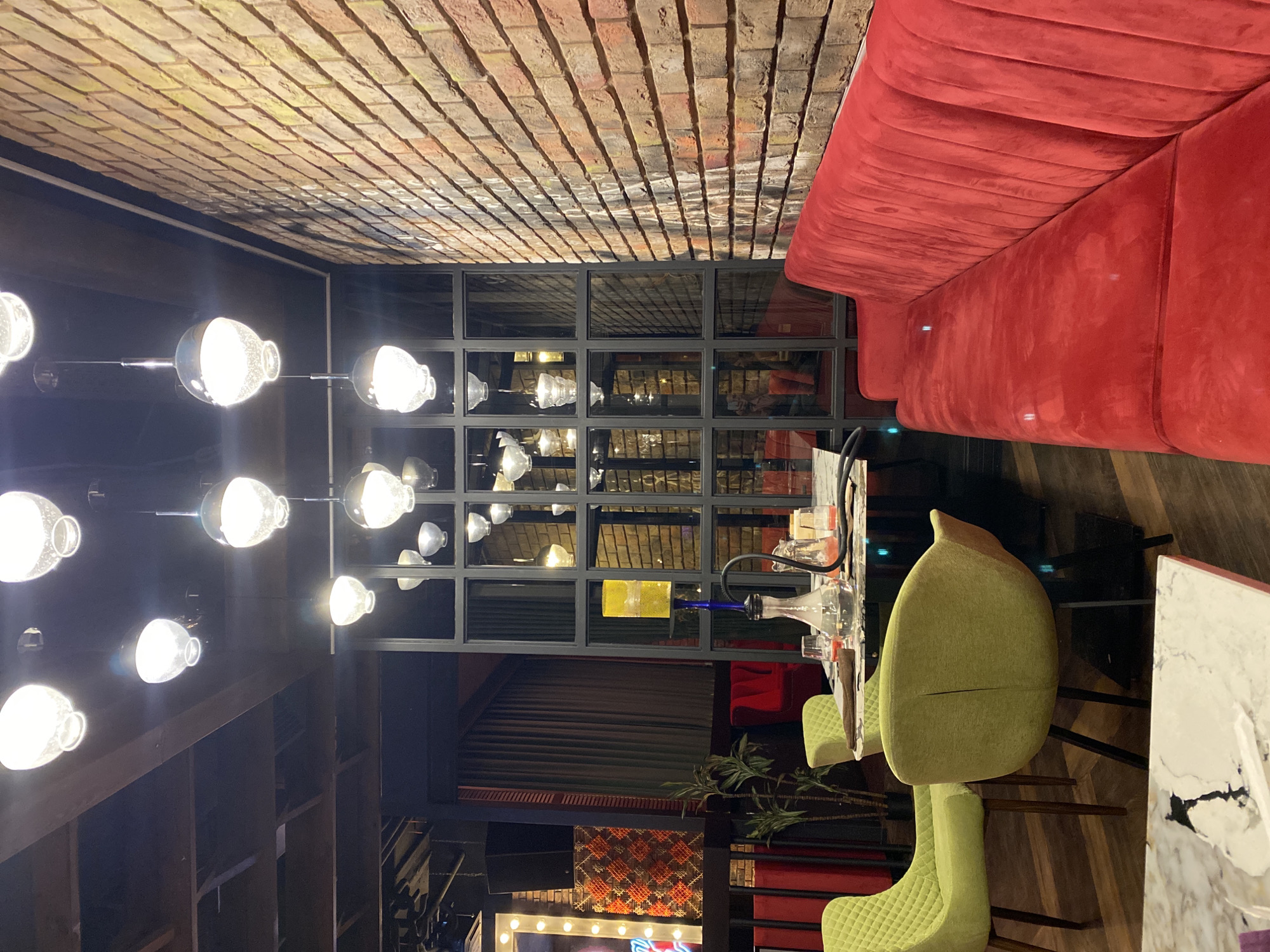 Here is a description of the image:
The image is an interior shot of a bar or restaurant. The ceiling is made of brick. On the left, there are multiple light fixtures with large, round bulbs hanging down. There is a large red couch along the top right side of the frame. Below the couch, there is a partition made of square glass windows. In the foreground, there is a table with a hookah and green chairs. There is a plant behind the chairs. In the background, there is a patterned wall and a lighted sign. The location is likely Yekaterinburg, Russia, based on the prompt.
