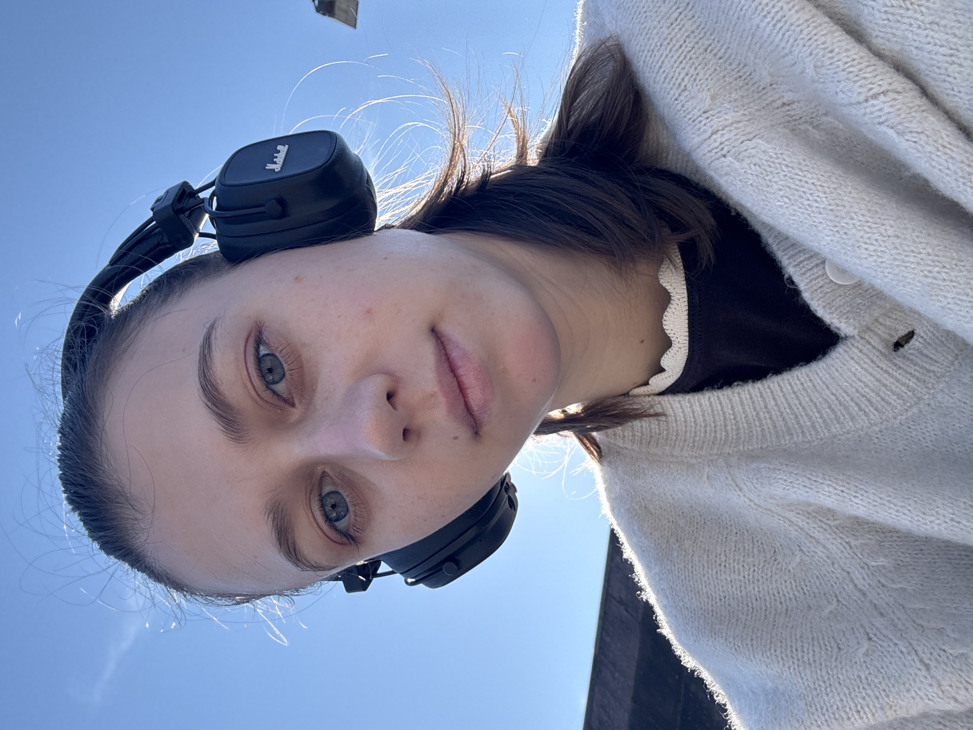 A young woman with light skin, blue eyes, and dark brown hair tied back is shown in a close-up, low-angle selfie. She is wearing black over-ear headphones with the "Marshall" logo visible. Her hair is slightly windswept, with strands catching the sunlight. She is dressed in a light-colored, knitted cardigan over a dark top with a ruffled collar. The background is a clear, bright blue sky. The image appears to be taken outdoors on a sunny day. There are no identifiable landmarks within the city of Miltenberg, Germany, visible in this image.