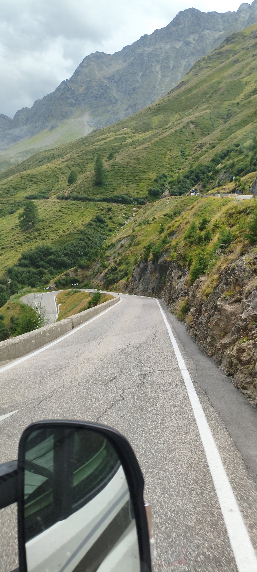 The image captures a winding mountain road with steep, green slopes covered in sparse vegetation and rocky outcrops. The road itself is paved and shows signs of wear with visible cracks. A white line marks the right edge of the lane. In the foreground, a black side mirror of a vehicle reflects the road and the white vehicle it is attached to. The sky is overcast and cloudy. The scene appears to be a scenic mountain pass in a rural or alpine setting, possibly the Timmelsjoch (Passo del Rombo), which is a high mountain pass in the Ötztal Alps connecting South Tyrol, Italy, and North Tyrol, Austria. The road is characteristic of alpine passes. There are no people visible in the scene.