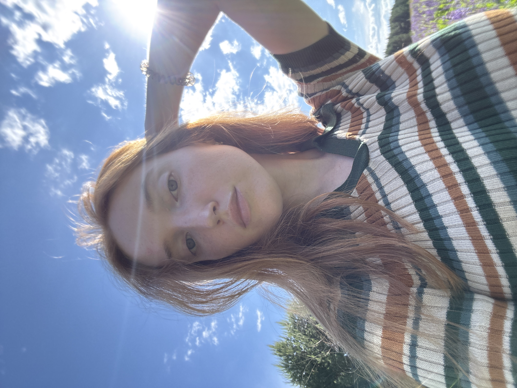 The image depicts a female individual outdoors under a bright blue sky with scattered white cumulus clouds. The sun is positioned in the upper right, generating a prominent lens flare. The individual has long, reddish-brown hair, fair skin with visible freckles on her nose and cheeks, and light-colored eyes. Her expression is neutral, and her gaze is directed straight at the camera. She is wearing a ribbed knit top with horizontal stripes in dark green, light green/teal, brown, and off-white, featuring a dark green V-neck collar. Her left arm is raised, with her hand positioned behind her head. A multi-beaded bracelet is visible on her left wrist. In the background to the left, green coniferous trees are visible. To the right, a field of purple flowers, consistent with lupines, extends towards a distant treeline. The scene is situated in the city of Chaikovsky, Russia.