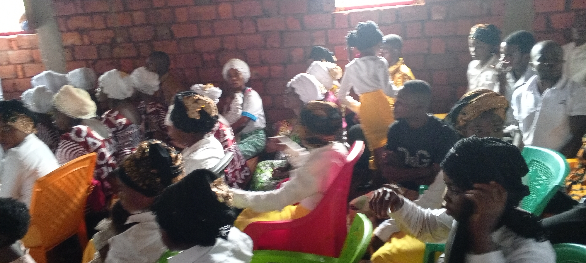 The image depicts a gathering of people in what appears to be an indoor setting, likely in Ngalula, Democratic Republic of the Congo. A brick wall is visible in the background. Most of the individuals are seated on plastic chairs. Many of the people are wearing white garments or head coverings, suggesting a religious or ceremonial event. One child is wearing a white top and yellow skirt. The general scene suggests a communal gathering or celebration.