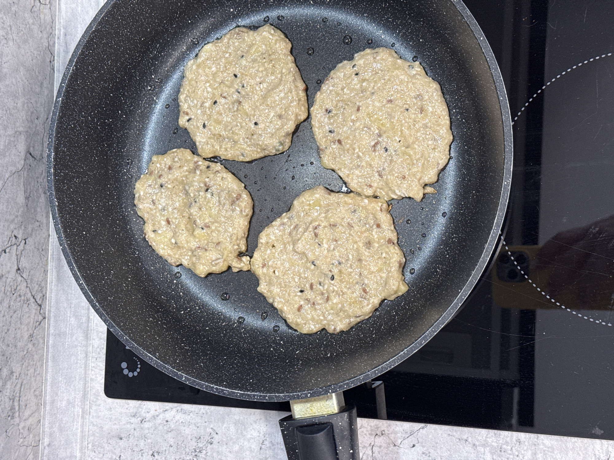 A top-down view shows four beige-colored patties cooking in a speckled black frying pan. Small bubbles of oil surround the patties. The frying pan is on a black ceramic stovetop. To the left of the stovetop is a light gray countertop. The location is unknown.