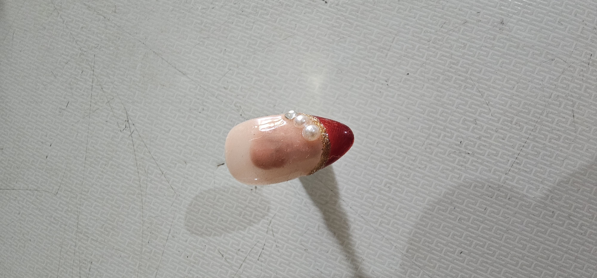 A single artificial fingernail, decorated with red and gold glitter at the tip and small pearls and rhinestones near the cuticle, lies on a light-colored surface with a subtle geometric pattern. The fingernail is pale pink with a reddish blush on the surface and appears to be made of acrylic or gel. A soft shadow is cast to the left of the fingernail. The surface is textured with a repeating maze-like pattern. The image is shot from a top-down perspective with shallow depth of field. The location in Puncak Jaya, Indonesia, is not discernible from the image.