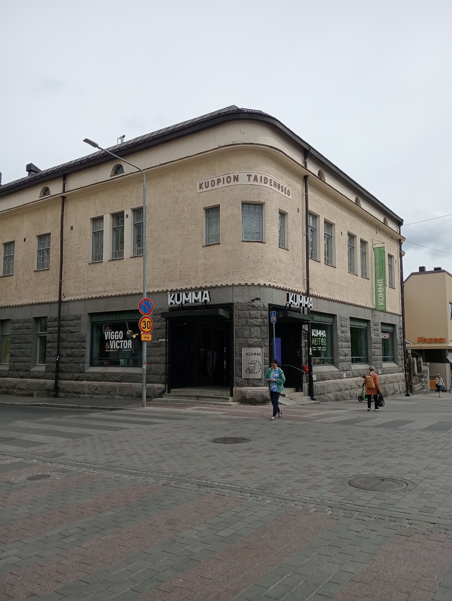 The image shows a street scene in Kuopio, Finland. The main subject is a pale yellow building with a curved corner, identified by text on its facade as "KUOPION TAIDEMUSEO" (Kuopio Art Museum). The building has a stone base and several windows on the upper floors. Multiple signs on the building indicate it houses the "KUMMA" art museum. One sign above an entrance reads "KUMMA" in large letters. Another green banner hanging from the building also says "KUMMA".
At street level, there are storefronts, including one with a sign for "VIGGO & VICTOR". There are also signs for "KUMMA AETIOS" and "GALERIA" on the ground floor. The street in front of the building is paved with interlocking stones. A pedestrian crossing is visible.
There are three individuals in the image. One person, wearing a blue and green jacket, is walking on the sidewalk in front of the building. Another person, with a backpack and carrying bags, is walking with a green electric scooter on the sidewalk. A third person is walking in the distance on the right side of the frame. There are also traffic signs, including a speed limit sign (30) and a prohibition sign. The sky is overcast.