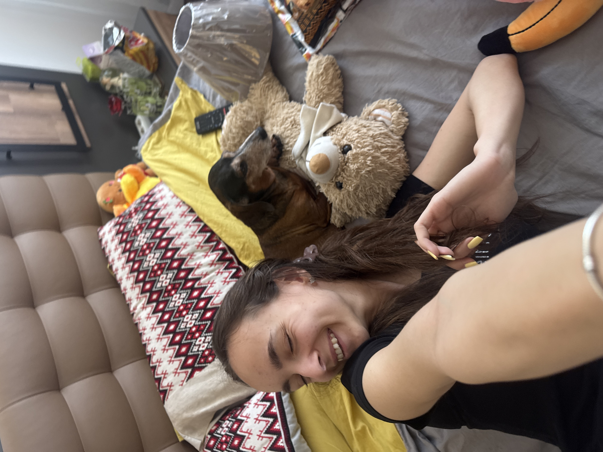 A young woman is taking a selfie indoors. She is lying on her left side on a bed with a brown tufted headboard. The bed has a gray sheet, a yellow blanket, and a decorative pillow with a red and white geometric pattern. A brown dachshund is lying on top of a stuffed teddy bear. A lamp, various toys, and other items are scattered on the bed. The woman is smiling and looking at the camera. Her fingernails are painted yellow. The setting seems to be a residential space in an unknown city. There is no information indicating that this image was taken in Sofia, Bulgaria.