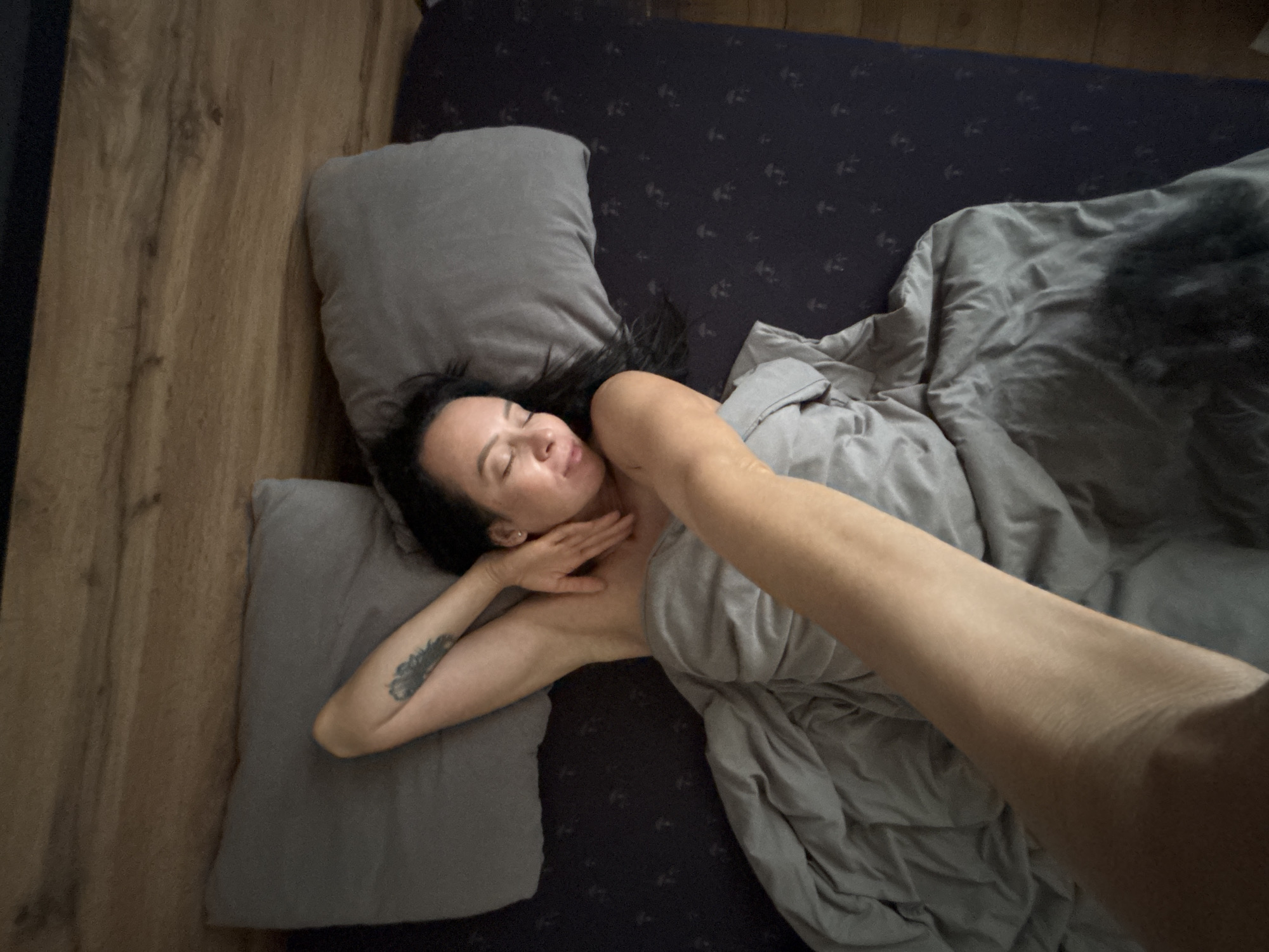 A woman is lying in a bed with gray sheets and pillows. She is on her back, with her left arm bent and her hand near her neck. Her right arm is extended towards the viewer. The bed has a dark blue patterned fitted sheet. The scene is indoors, with a wood-paneled wall on the left. The location is unspecified; the city of Chaikovsky, Russia, is not discernible from the visual information.