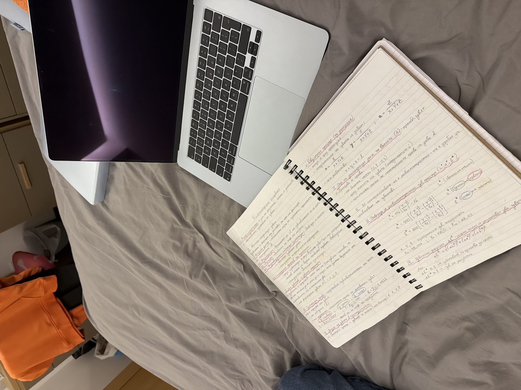An overhead shot captures a study or work setup on a light grey duvet cover, likely in a bedroom in Sofia, Bulgaria. A silver MacBook laptop is open on the left, with its screen off or displaying a dark reflection, and its keyboard visible. To the right of the laptop is a spiral-bound notebook filled with detailed handwritten notes in Bulgarian.
The notes cover topics related to color theory and physics, including "Калориметрични измервания" (Colorimetric measurements), "Фактори определящи цвета" (Factors determining color), and "Двумерно простр. (ху диаграма)" (Two-dimensional space (xy diagram)). Formulas and equations involving X, Y, Z coordinates, and the L*a*b* color space are prominent, along with terms like "Цветна разлика" (Color difference). Some headings and words are highlighted in pink and green ink, indicating emphasis.
In the bottom left corner, part of an orange hoodie is visible, and a glimpse of a blue denim-like fabric can be seen near the bottom right. The overall impression is a focused academic environment set in a comfortable, personal space. The lighting is bright and even, suggesting either daytime or good artificial illumination.