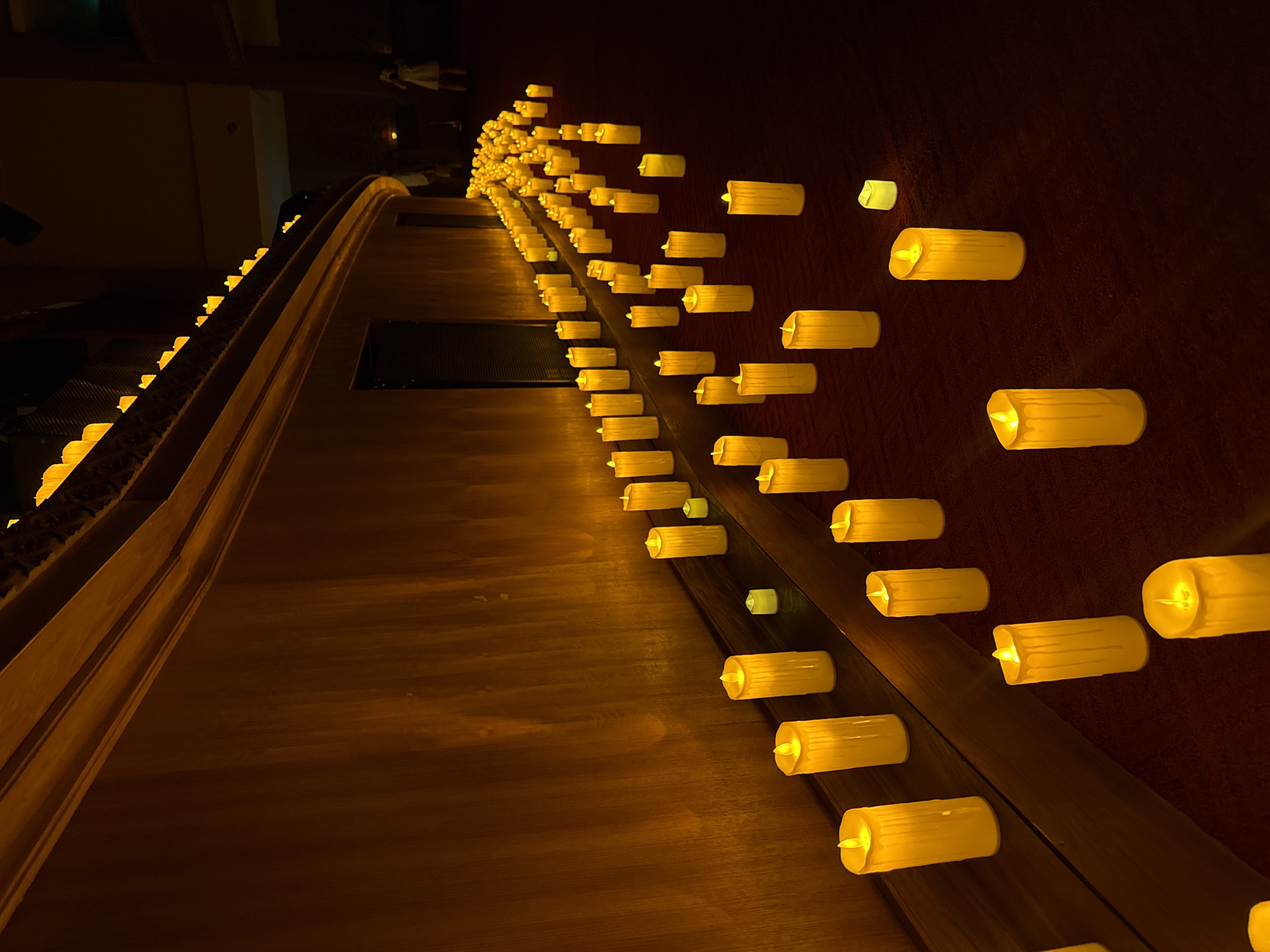 A long row of cylindrical, artificial candles with yellow lights line a dark brown wall. The row extends into the distance, decreasing in size. The wall is adjacent to a lighter brown surface. The location is North Kuta, Indonesia.