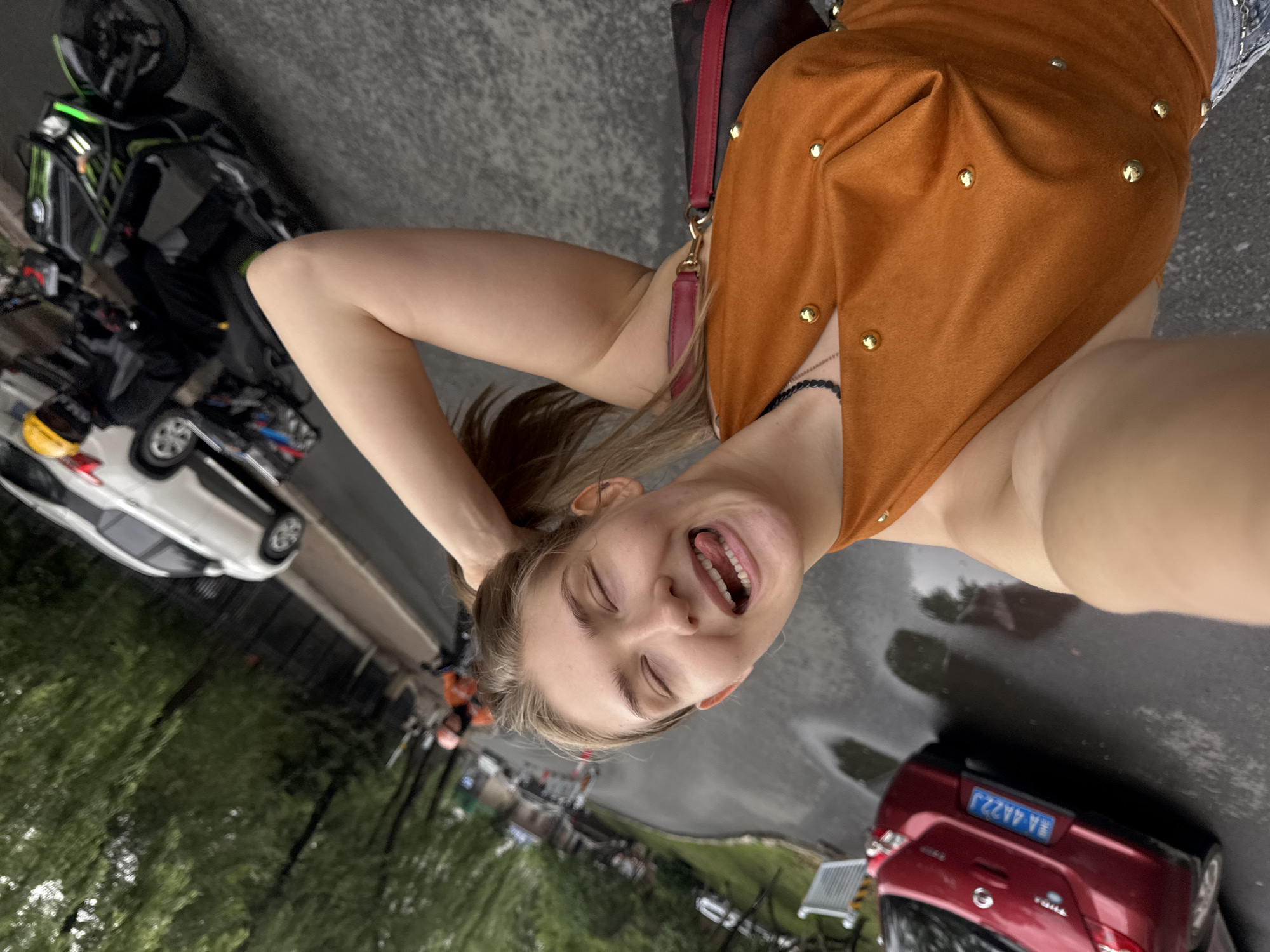A young woman with blonde hair pulled into a ponytail is taking a selfie outdoors. She is wearing an orange halter top adorned with gold studs and has her right arm raised, with her hand in her hair. Her eyes are closed, and her tongue is sticking out. She is standing on a wet asphalt road with puddles reflecting the overcast sky. In the background, a red car with a blue license plate reading "A 4A22J" is parked on the left side of the road. Further down the road, a white SUV is driving, and behind it, a person in black riding a green motorcycle is visible. Trees line the left side of the road, and a black metal fence runs parallel to the street. The scene appears to be a street in Xiangfang District, China, on a rainy day.