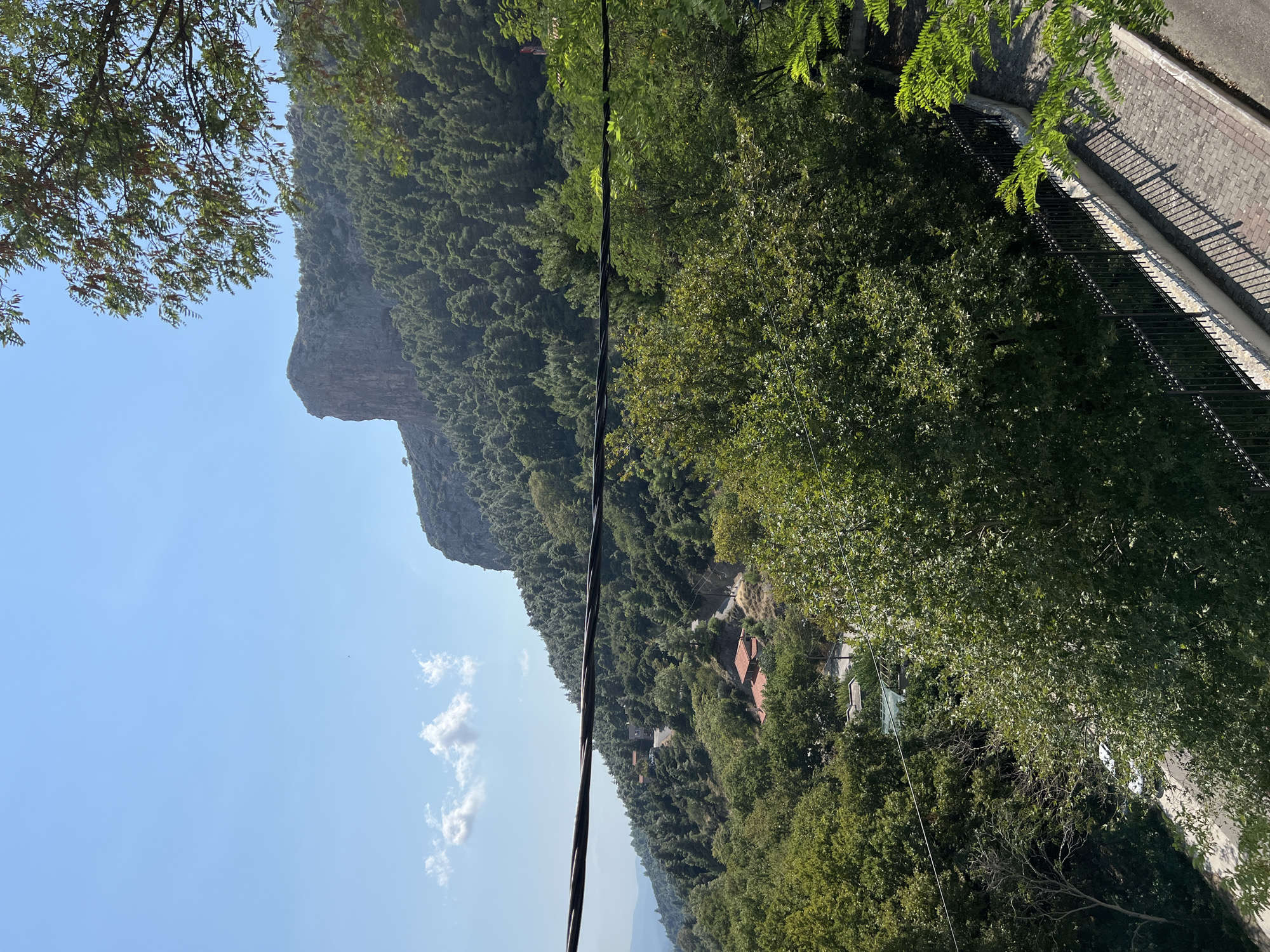 The image depicts a scenic view of a mountainous landscape near the village of Stení Dirfýos, Greece. In the foreground, a paved walkway with a black metal fence runs along the right side. Beyond the fence, lush green trees dominate the scene, with their branches obscuring parts of the view. Several power lines are visible, stretching across the middle ground of the image.
Further back, the terrain slopes upwards, covered in dense evergreen forest. Nestled within the trees are a few scattered red-roofed buildings, indicating a small settlement or individual dwellings. A winding road is visible, snaking through the hillside and connecting these structures.
In the background, a prominent, sheer rock face rises majestically above the forested mountain. The sky is clear and blue, with a few scattered white clouds. The overall impression is one of a serene and natural environment, likely a rural or semi-rural area on a mountainside.