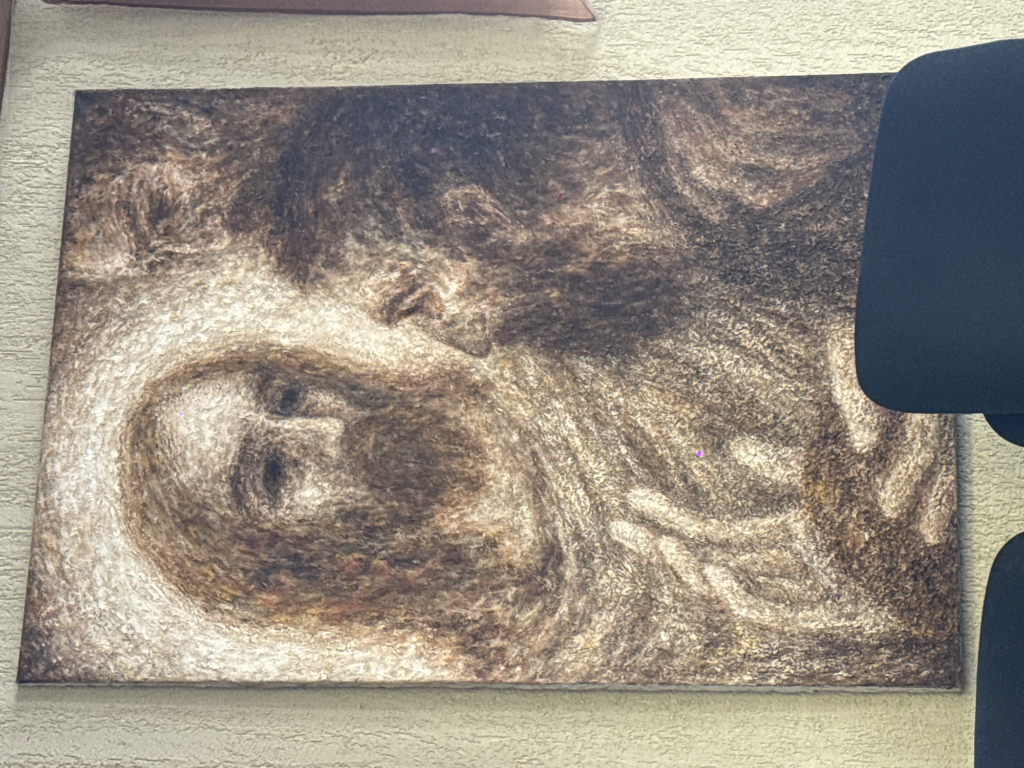 The image depicts a textured artwork hanging on a light-colored wall. The artwork appears to be an abstract or stylized portrait of a face rendered in shades of brown and white. The person's face, or at least its suggestion, is centrally located on the left half of the canvas, while the top and right sides of the artwork feature blurred and textured patterns. A black chair is partially visible on the right side of the image, obscuring part of the artwork.
There is no indication of location in the city of Moscow, Russia.