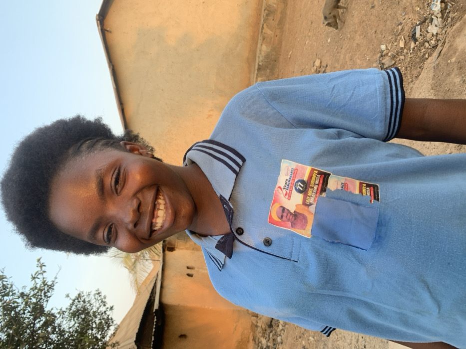 A smiling girl stands outdoors, wearing a light blue polo shirt with dark blue trim. Patches, one featuring a man's portrait and the text "2 Years Remembrance" are affixed to her shirt. A building is partially visible in the background, and a primate can be seen at the top of the image. The photo appears to have been taken during daylight hours, with natural light illuminating the scene.