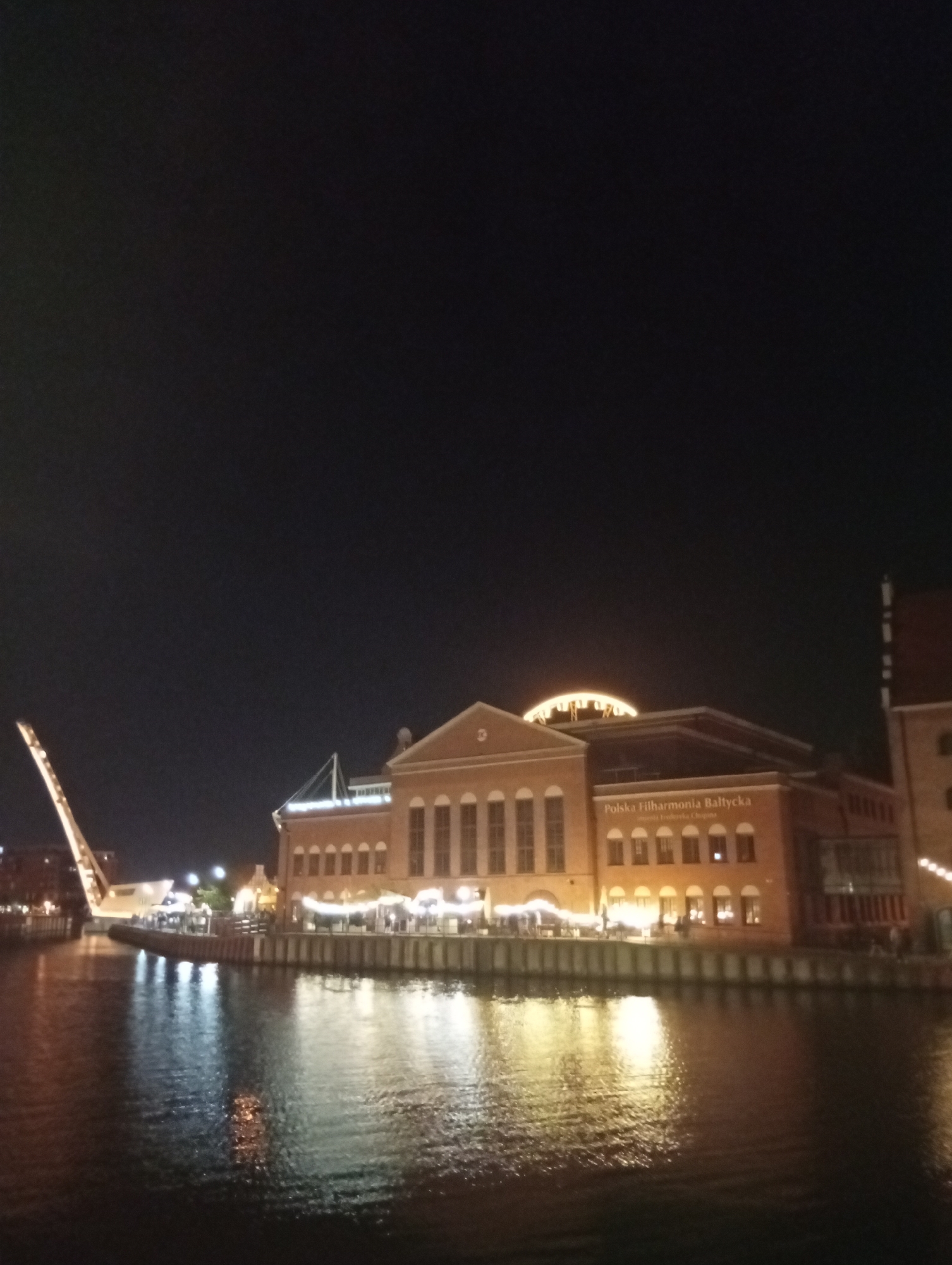The image depicts the Polska Filharmonia Bałtycka (Baltic Philharmonic) building situated on the Motława River in Gdańsk, Poland. It is nighttime, and the building is illuminated, with lights reflecting on the dark water. The architecture of the philharmonic hall is visible, featuring a large, light-colored dome structure on top. The building's facade is made of brick, with numerous windows and arched entrances. Along the waterfront in front of the building, there are outdoor seating areas with lights, suggesting a restaurant or cafe. To the left, a large, modern bridge structure, possibly a bascule bridge, is partially visible, extending upwards at an angle. The sky is dark, indicating it is night. The overall scene is one of urban nocturnal ambiance by a waterfront.