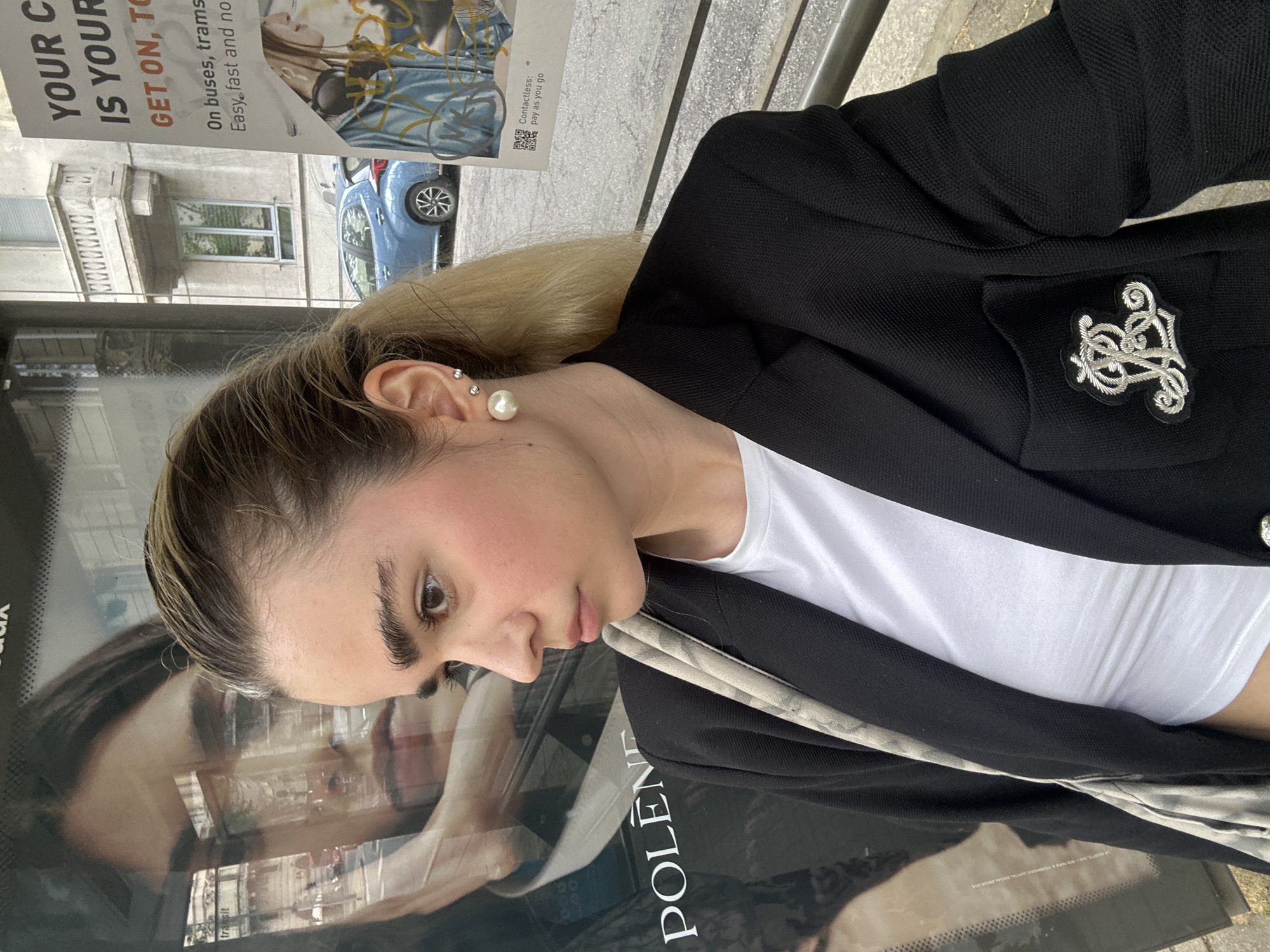 A portrait-style photograph shows a fair-skinned woman with light brown hair, who is standing in front of a bus stop shelter that displays an advertisement for the "Polène" brand. Her clothing consists of a white shirt, and a black blazer, with an ornamental silver embellishment. The woman is wearing pearl earrings and has other piercings. The advertisement for public transit is visible, with the words "YOUR C IS YOUR GET ON, TO" and a partial phrase indicating payment options. In the background, a blue car and buildings are visible. Based on the visual evidence, this scene is plausibly set in Milan, Italy, given the brand of the advertisement is Italian.