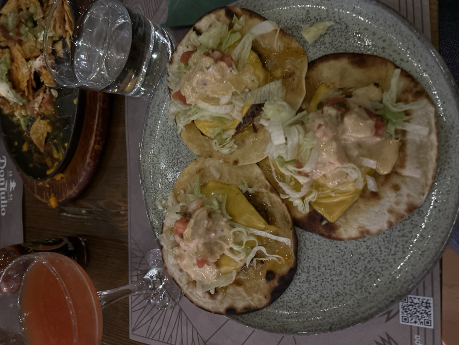 An overhead shot captures a dining scene, likely in a restaurant in Melissia, Greece, featuring several dishes and drinks on a wooden table. The primary focus is a speckled, light grey ceramic plate holding three soft tacos. Each taco is generously filled with shredded lettuce, diced red tomatoes, a creamy light-colored sauce, and visible chunks of what appears to be melted yellow cheese or a yellow fruit/vegetable, possibly pineapple or bell pepper. One taco also reveals a dark filling, likely meat. The tacos have slightly charred edges on their tortillas, suggesting they were warmed.
To the left, a partially eaten serving of nachos rests on a rustic wooden serving board with a dark metal base. Remnants of tortilla chips, melted cheese, and other toppings are visible. A clear glass filled with water is positioned between the nachos and the tacos. In the lower left foreground, a stemmed glass holds a vibrant reddish-orange cocktail.
A white placemat or tablecloth with a subtle geometric pattern is visible under the plates and drinks. Visible text includes "Don Julio" printed on a napkin or paper placed under the nachos board, and a QR code is clearly visible on the placemat in the bottom right corner. The lighting appears to be indoor ambient light, creating reflections on the glassware. The overall scene suggests a casual meal or snack.