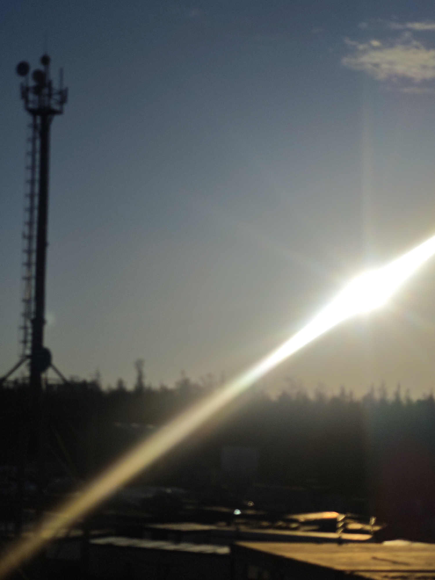 A silhouette of a telecommunications tower stands against a blue sky, partially obscured by a bright, angled sunbeam. The tower has multiple satellite dishes and a ladder. The foreground is dark, appearing to be an indistinct structure. A line of trees is visible behind the foreground structure. The location is possibly in Unknown, Russia, though there is no definitive information in the photo to confirm this.