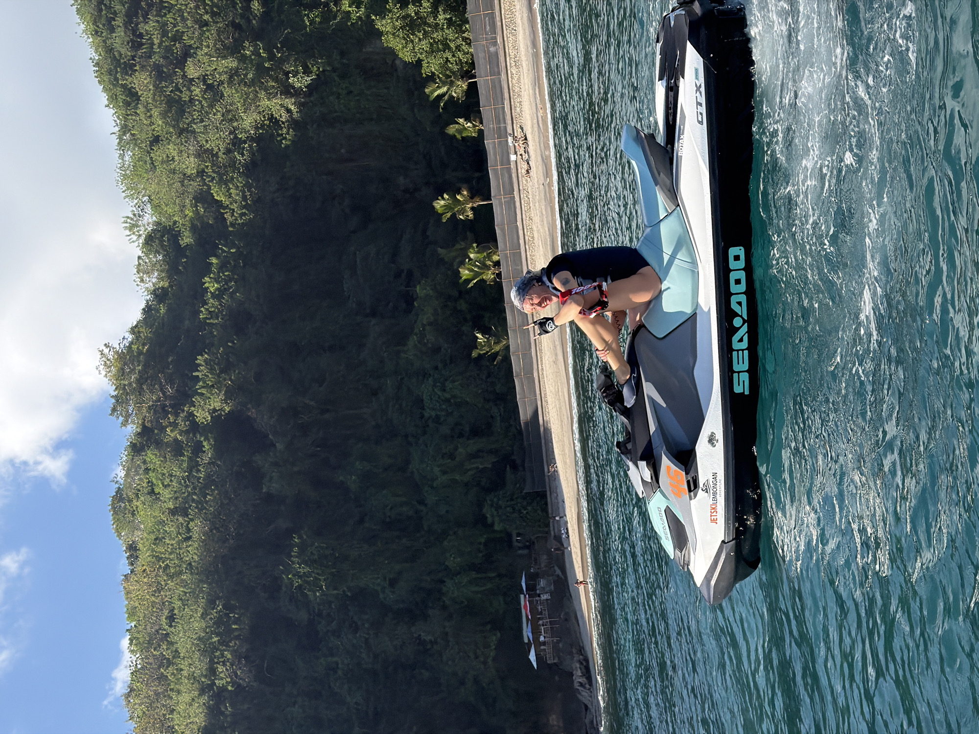 A person rides a Sea-Doo jet ski in the turquoise waters of Nusa Penida, Indonesia, with a lush green hillside and a sandy beach in the background. The person, wearing a dark blue rash guard and patterned shorts, sits on the jet ski, looking towards the camera with a wide smile and a bandana on their head. They have their left arm raised with fingers pointed. The jet ski, a Sea-Doo GTX model, is white with grey and light blue accents and features the text "JET SKI LEMBONGAN ADVENTURE" on its side. Behind the jet ski, a narrow sandy beach is visible, lined with palm trees and a small building with colorful umbrellas. Further back, the densely vegetated hillside rises steeply, covered in various shades of green trees. The sky above is blue with scattered white clouds. The water around the jet ski is rippling, creating white foam and streaks.