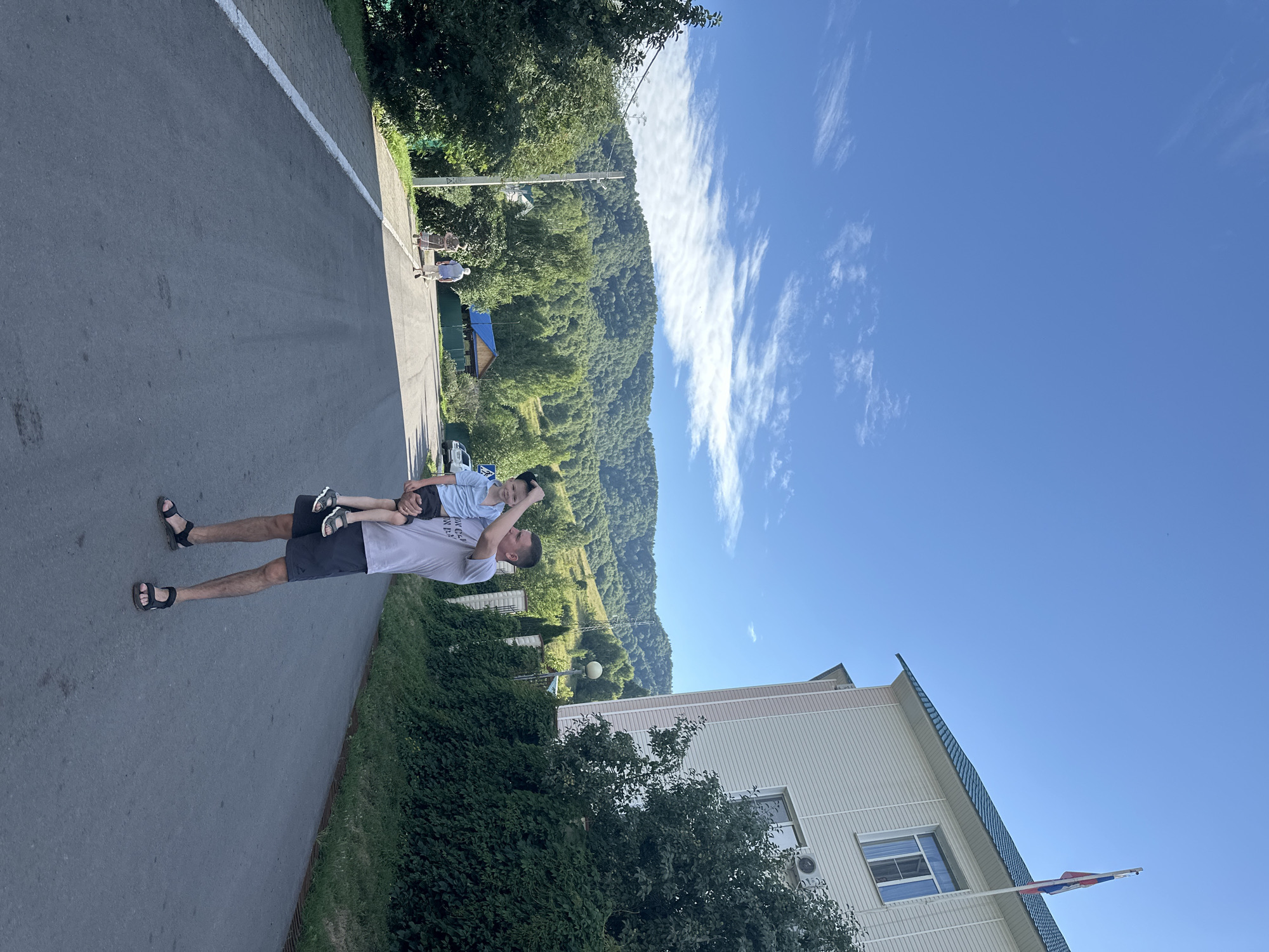 The image depicts a scene on a paved road in Danilovka, Altai Krai, Russia. In the foreground, a man is holding a young child. The man is wearing a light-colored t-shirt and dark shorts with black sandals. The child, dressed in a blue shirt and dark shorts with black sandals, is being held in the man's left arm, with their right arm raised. To the right of the road, a multi-story building with a light-colored facade and a green roof stands, flying a Russian flag from a flagpole. Lush green trees and bushes surround the building and line the sides of the road. In the background, rolling hills covered in dense green forest rise under a clear blue sky with scattered white clouds. Further down the road, two figures are walking away from the camera, and a white car is parked on the right side. The overall atmosphere suggests a bright, sunny day in a rural or semi-rural setting.