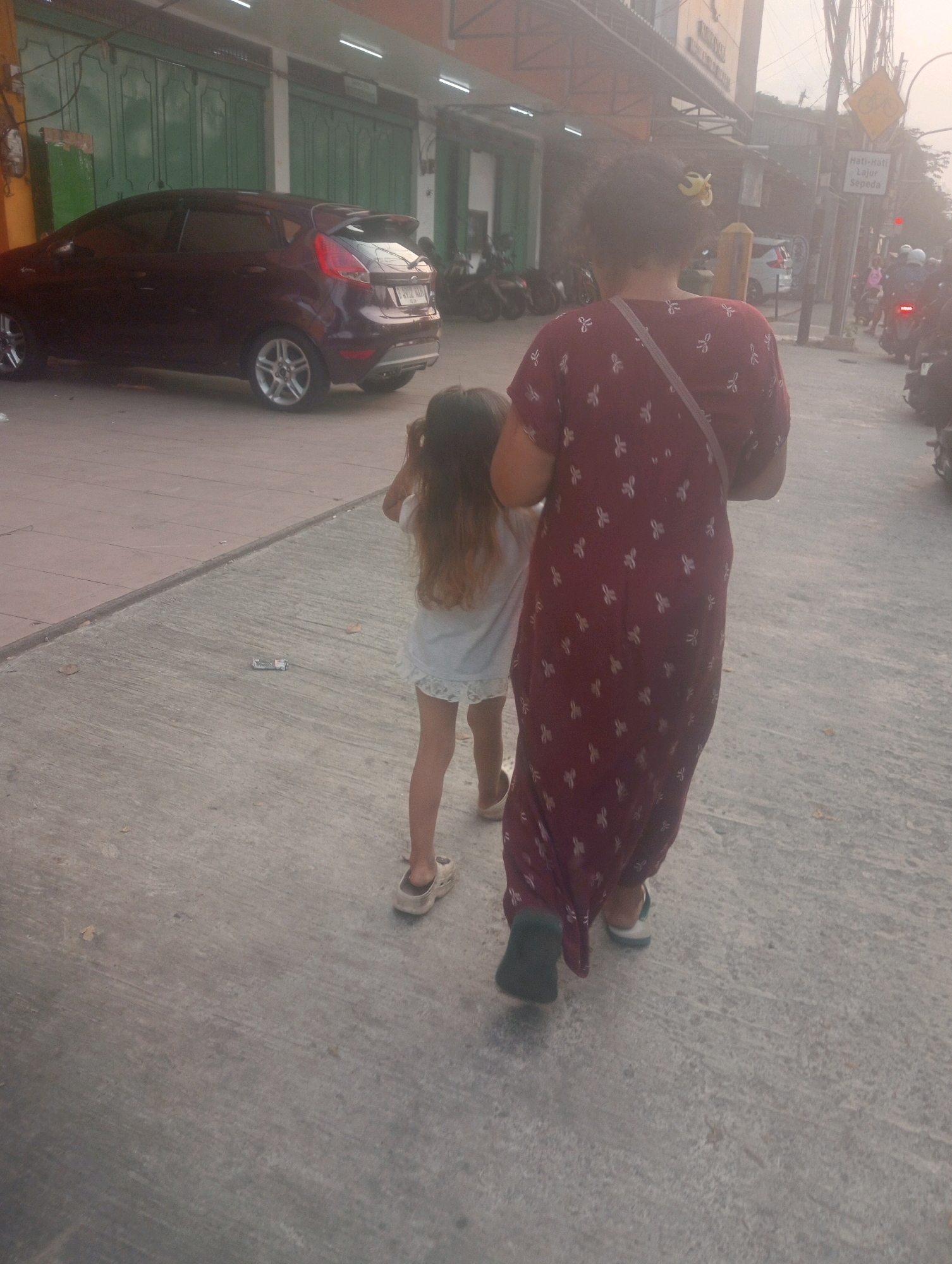 A woman and a young girl walk on a sidewalk in front of buildings. The woman is wearing a long, maroon dress with white floral patterns and a beige strap across her chest. The girl is wearing a short, light-colored dress and has long, brown hair. They are walking away from the camera. In the background, there is a dark-colored car parked on the street, and several motorcycles are also visible. Buildings with green doors line the street. A yellow traffic sign with a bicycle symbol is also present, along with a red traffic light. The sky is hazy, suggesting an overcast or dusty atmosphere. The scene appears to be in an urban environment, likely a commercial area, in East Jakarta, Indonesia.