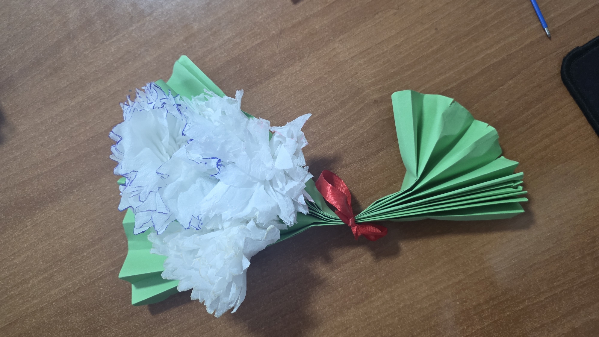 A handmade bouquet of three white paper flowers with blue-outlined edges and green paper leaves is displayed on a wooden surface. The bouquet is tied with a red ribbon. The wooden surface appears to be a table. The lighting suggests an indoor setting. There is no indication of the city of Tokmok, Kyrgyzstan, or any specific location within it.
