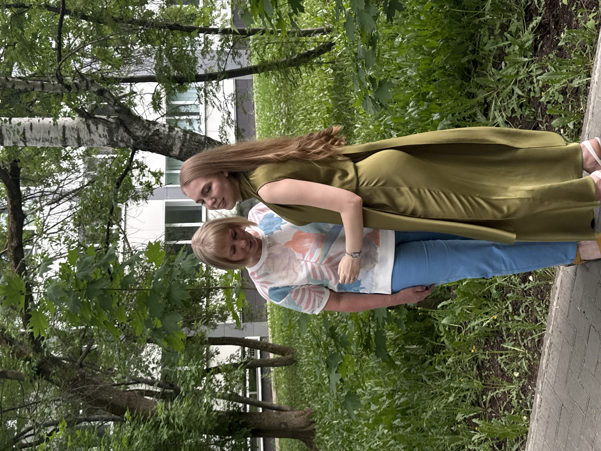 The image depicts two women outdoors in what appears to be a park-like area, possibly in Perm, Russia. The younger woman is wearing a long, green dress and the older woman is in a white t-shirt with a floral pattern and blue pants. They are standing close to each other, possibly embracing. The setting includes greenery, trees, and a building in the background. The photo is taken at ground level.