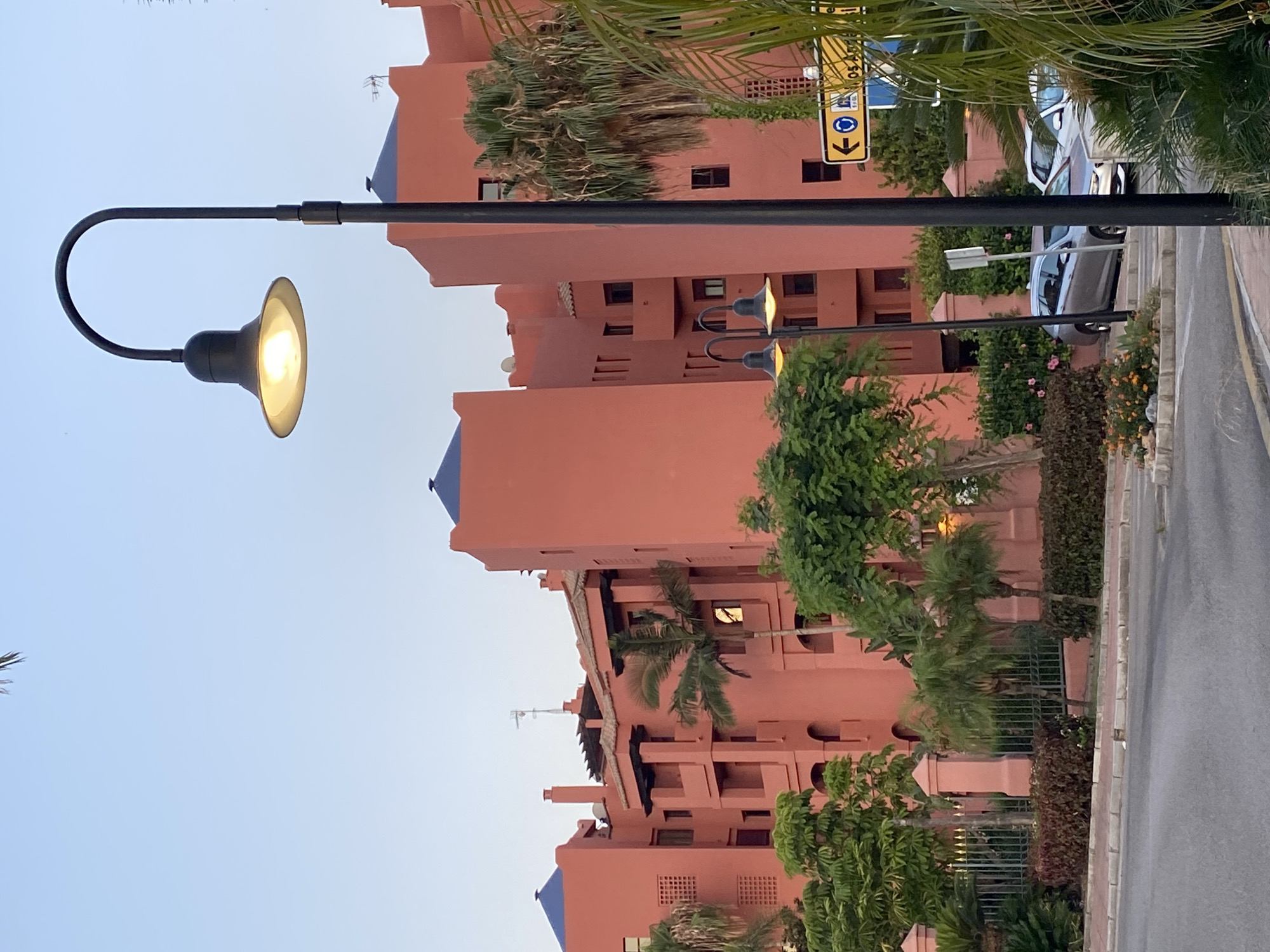 The image shows a street scene in Estepona, Spain, in the late afternoon or early evening. The main subject is a large, coral-colored building with blue-tiled roofs, characteristic of Mediterranean architecture. Several palm trees and other lush greenery surround the building. A street lamp with its light on is prominently featured in the foreground. In the background, a street sign is visible, indicating directions. A few cars are parked along the street. The sky is a clear, pale blue, suggesting a pleasant climate.