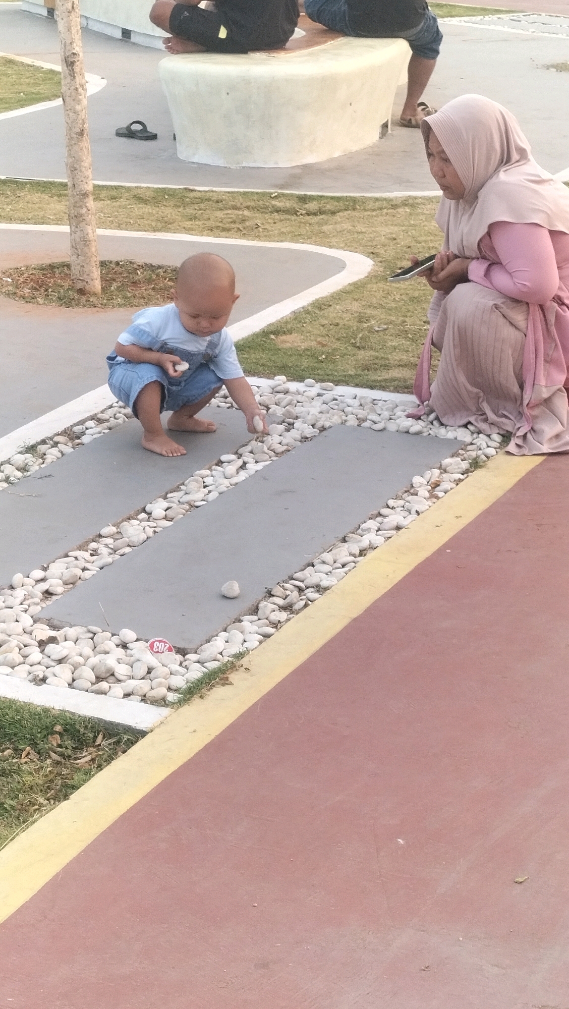 An overhead shot shows a child, wearing a light blue t-shirt and denim overalls, crouching on a paved area near a patch of grass. The child is surrounded by small white stones and is holding a white stone in their right hand, with their left hand extended towards the ground. To the child's right, a woman wearing a light pink hijab and a matching pink dress is kneeling and holding a mobile phone in her hands. In the background, there is a light-colored, curved concrete bench, and a tree trunk is visible to the left of the child. The ground is a mix of grass and pavement with a yellow line marking the edge of the red paved area in the foreground. The scene appears to be an outdoor public space, possibly a park, in Ketanggan, Indonesia.