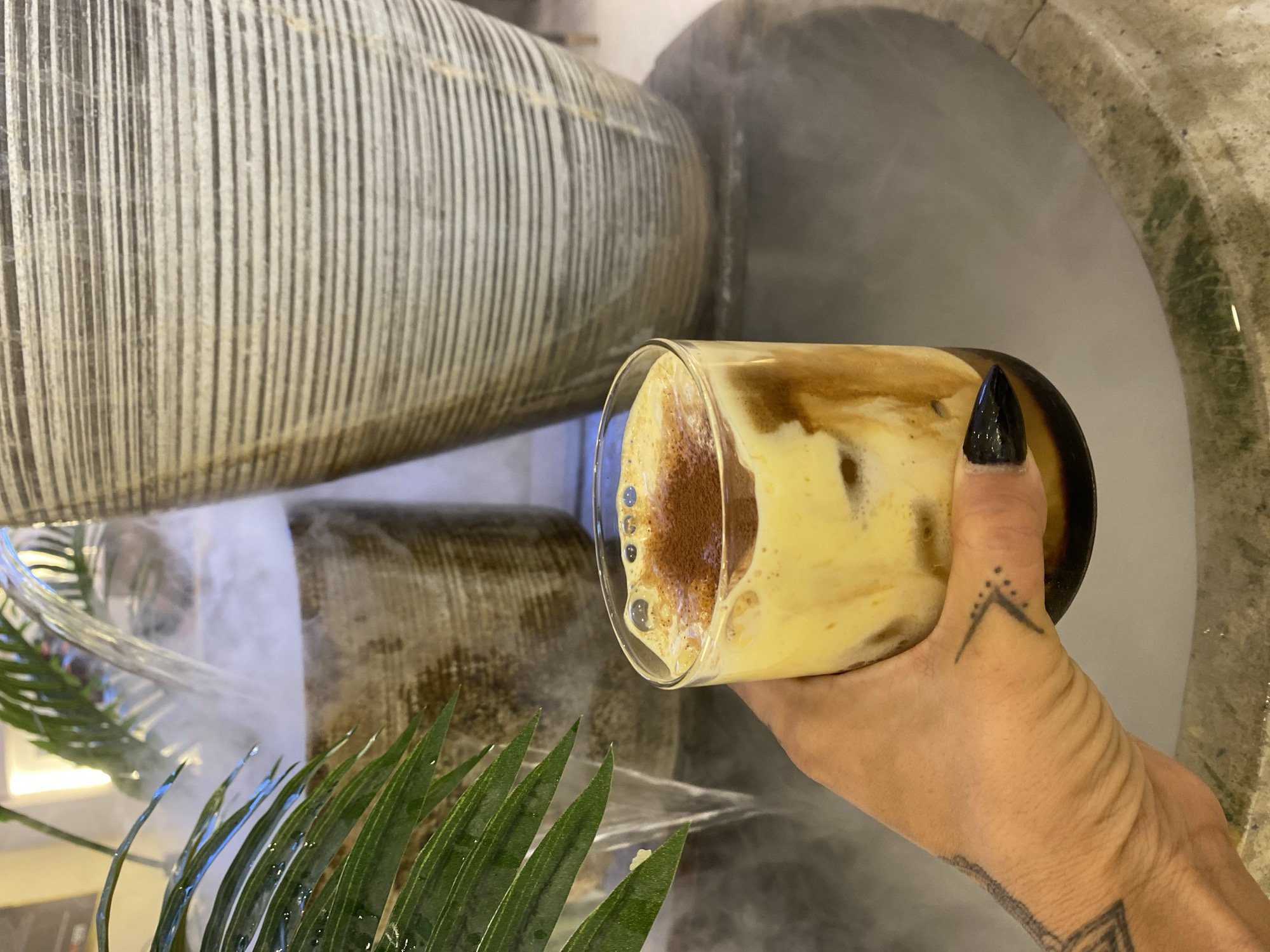 A hand with black fingernails holds a glass of layered beverage, possibly coffee, in front of a decorative fountain or water feature. The fountain consists of stacked, textured cylinders with water flowing around them, creating a misty effect. Green leaves of a plant are visible at the bottom left of the frame. The location is Phu Thuy Ward, Vietnam.