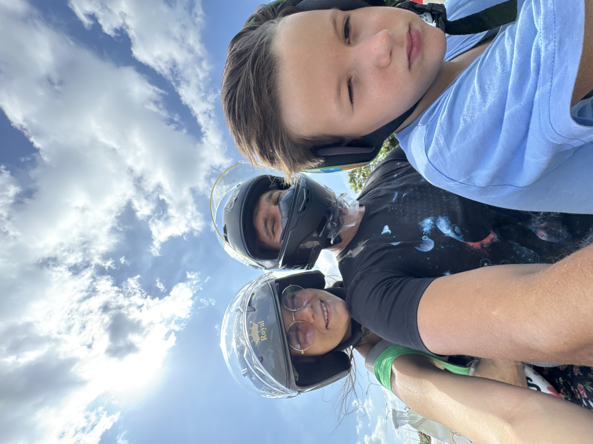 A close-up, slightly low-angle photograph captures three individuals on a bright, sunny day, presumably on a moving motorcycle or scooter in Mui Ne, Vietnam. The upper two-thirds of the frame are dominated by a clear blue sky interspersed with scattered white cumulus clouds. A strong sun flare emanates from the upper-left corner, indicating direct sunlight.
From left to right, the subjects are:
1. A woman, appearing in her 20s or 30s, is positioned in the foreground. She wears a dark grey open-face helmet with a clear visor, adorned with a gold "Royal" brand logo and crown emblem. Round, gold-rimmed sunglasses with pink-tinted lenses cover her eyes. She smiles directly towards the camera, with dark hair strands visible and wind-blown. A green strap, possibly from a bag or garment, is visible on her left shoulder. Her right forearm extends towards the camera.
2. A man, appearing in his 30s or 40s, is situated behind the woman. He wears a black full-face helmet with the clear visor lifted, revealing a chin strap and a light-colored neck or chin covering. His eyes are visible, looking forward. He wears a black short-sleeved t-shirt featuring a cosmic or galaxy-themed print with blue and purple patterns.
3. A boy, approximately 8-12 years old, is positioned to the right of the man. He wears a dark helmet with a visible chin strap and a colorful stripe (green, blue, red) on its side. His light brown hair is windswept, and his eyes are partially squinted, looking slightly downwards or away from the camera. He wears a light blue short-sleeved t-shirt.
All three individuals are wearing helmets, indicating their involvement in vehicular travel. The background is blurred, showing indistinct structures resembling utility poles or streetlights on the left and a suggestion of green foliage on the right, consistent with an outdoor roadside environment.