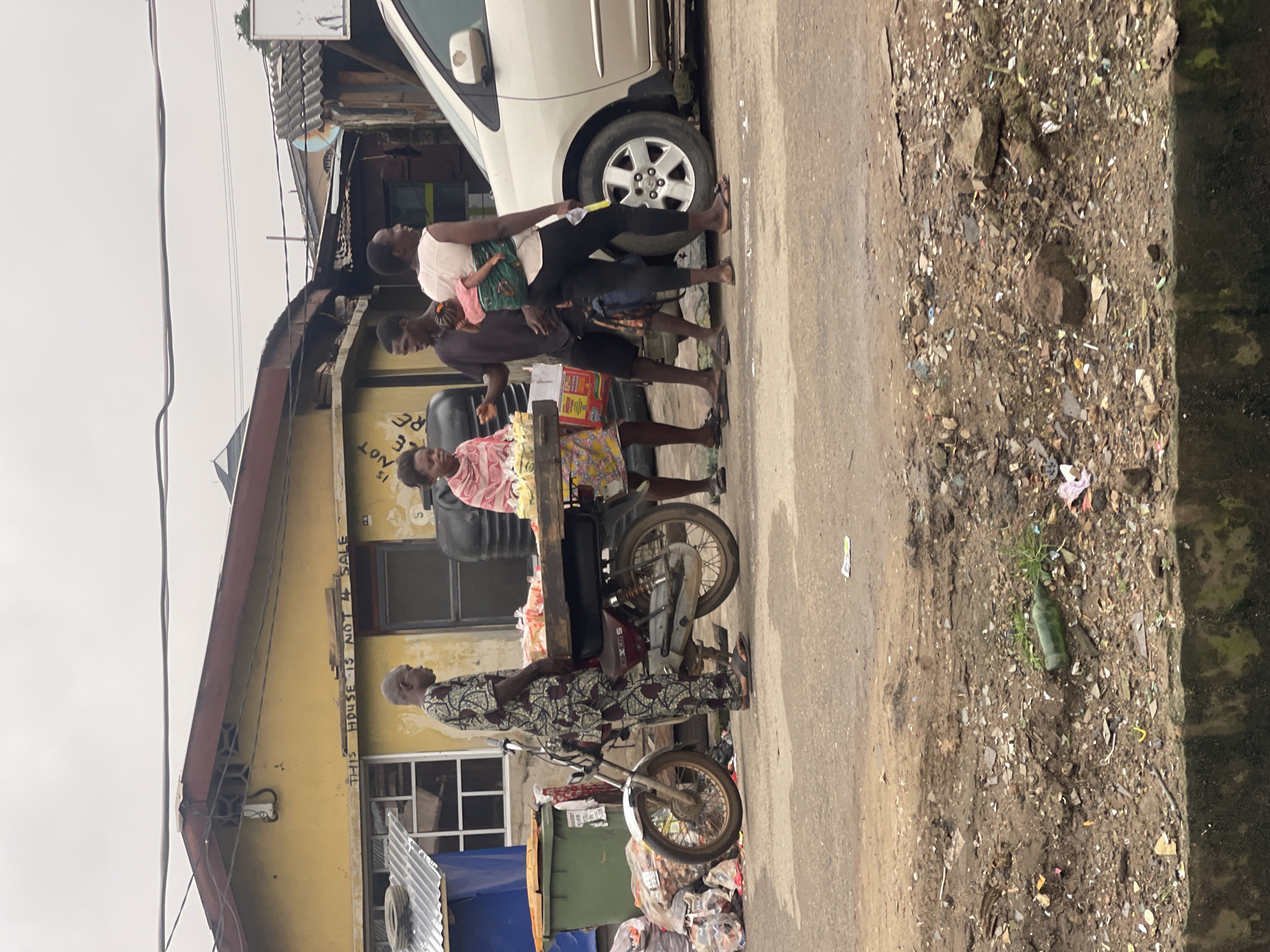 In Akowonjo, Nigeria, on an overcast day, a street scene unfolds. A man in a patterned garment stands beside a motorcycle with a wooden platform loaded with goods. To his right, a woman holding a baby on her back converses with another woman and a man standing near the motorcycle's platform. In the background, a yellow building with red trim bears the inscription "THIS HOUSE IS NOT FOR SALE". A silver car is parked to the right of the scene, and piles of debris are visible on the left and foreground. The ground is paved with asphalt, and the edges of the road are lined with dirt and discarded materials.