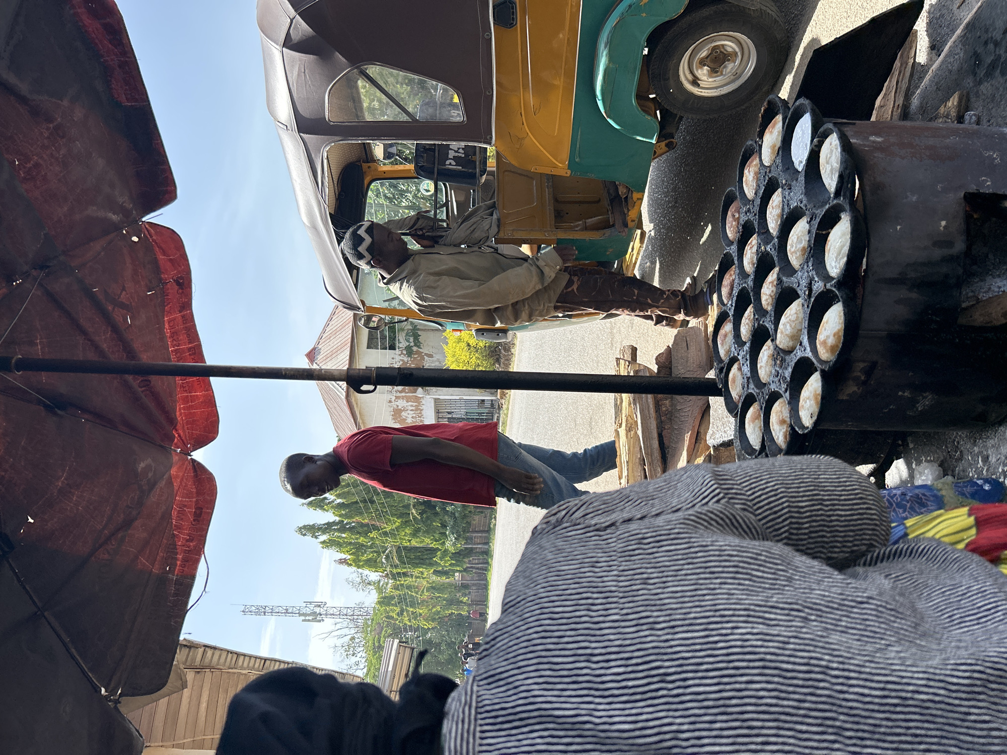 This photograph, taken in Jos, Nigeria, captures a street scene dominated by street food preparation and vehicular activity. An individual wearing a khaki-colored outfit and a cap is positioned near a yellow and green auto-rickshaw. Adjacent to this vehicle is a dark, metallic, multi-compartment cooking apparatus filled with small, circular dough-like items, suggestive of street food being prepared. Another person dressed in a red shirt and jeans is standing nearby. In the foreground, a gray striped garment is partially visible, obscuring the view. A weathered red umbrella provides shade over the scene. In the background, there are buildings and trees, contributing to the urban landscape of Jos.