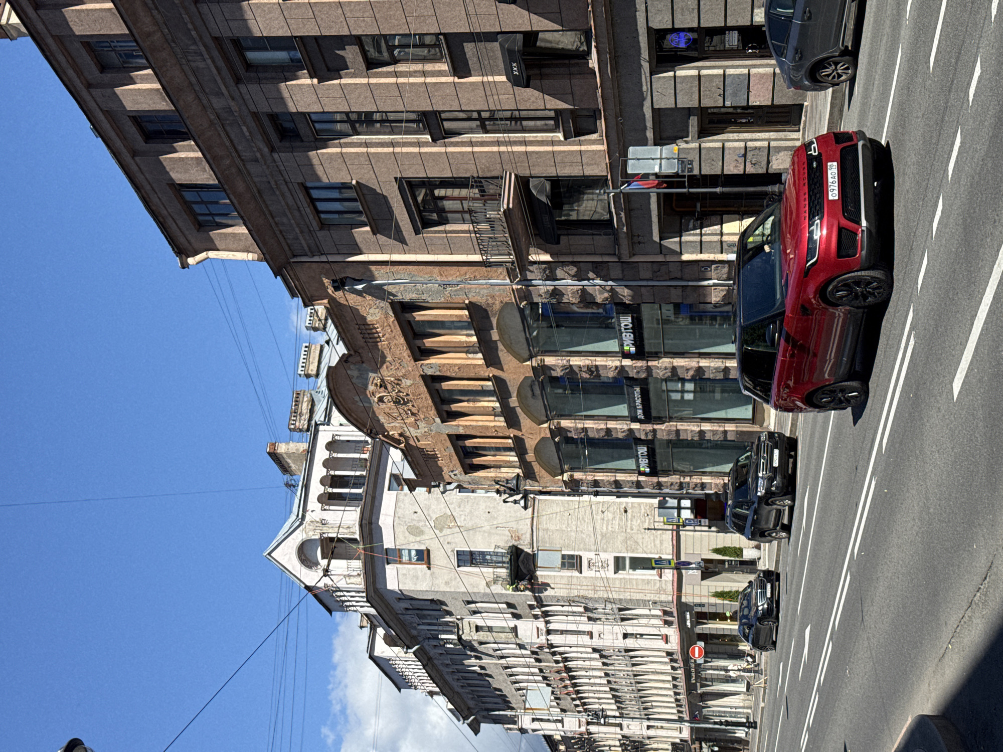 This image captures an urban street scene on a clear, sunny day, likely in the mid-morning or afternoon given the strong shadows. The perspective is slightly angled, looking up at multi-story buildings that line a paved road.
The buildings are a mix of architectural styles. The closest building on the left appears older, with ornate detailing and weathered stone facades. Further down the street, more modern buildings with large glass windows and commercial signage are visible. Several cars are present on the road, including a prominent red Range Rover in the foreground, and two black SUVs behind it. White lane markings are clearly visible on the asphalt.
There are a few people visible, including what appears to be a construction worker in a yellow vest on a balcony on one of the buildings. The overall atmosphere is one of everyday urban activity. The bright sunlight and clear blue sky suggest pleasant weather.
Visible text includes signage on the businesses, such as "ДОМ КРАСОТЫ" (House of Beauty) and "ФИВТОШ" on the glass storefronts. The license plate of the red Range Rover reads "0976 AO 78". The presence of Russian text on the signs and the license plate format suggest the location is likely in Russia.