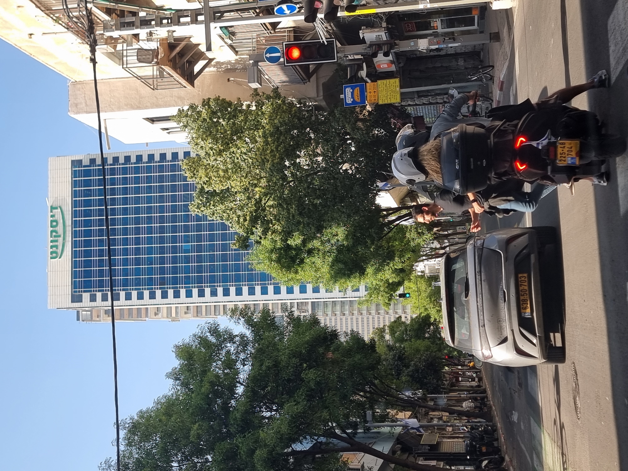The image is a street scene in Tel Aviv, Israel. A tall modern building with the Hebrew word "Discount" (דיסקונט) in green letters on its side dominates the background. In the foreground, a grey car and a motorcycle are on the street. The car has a license plate that reads "638:50-703" in Hebrew characters, while the motorcycle's license plate is "225-48-704". The motorcycle is being driven by two people, both wearing helmets. The street is lined with trees. A traffic light is visible, showing a red signal. The scene appears to be captured on a sunny day.