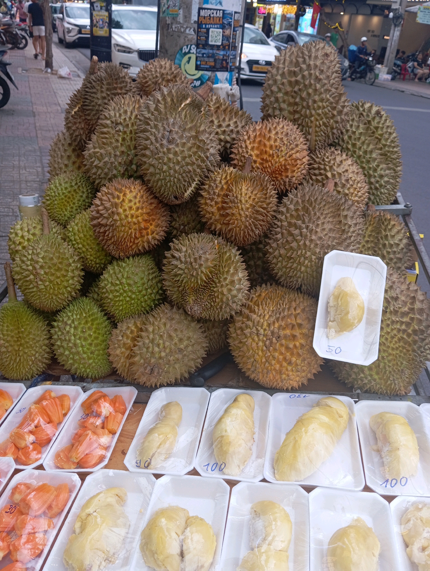 The image depicts a street scene in Nha Trang, Vietnam, featuring a market stall laden with durian fruits and other produce. In the foreground, rows of white styrofoam trays are arranged, containing pre-portioned servings of durian flesh. Some trays are labeled with prices, such as "50" and "100". To the left of the durian trays, there are also trays filled with pieces of jackfruit, also wrapped in plastic.
Dominating the upper half of the image is a large collection of whole durian fruits, piled high on a cart. The durians vary in color, from bright green to a more golden brown, and their characteristic spiky husks are clearly visible.
In the background, the urban environment of Nha Trang is evident. A street runs alongside the market stall, with various vehicles, including cars and motorcycles, visible. Pedestrians can be seen walking on the sidewalk, and shop fronts are present on the other side of the street. The overall impression is a bustling street market scene in a Vietnamese city.