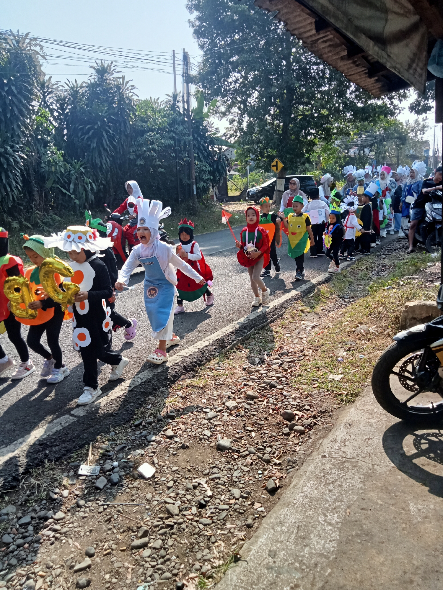 In Dororejo, Indonesia, a group of children in colorful costumes participates in a street parade under a bright, clear sky. The children are dressed as various fruits and vegetables, including a girl in a strawberry costume with a red headscarf, a girl in a corn costume, and a child dressed as a sunflower. Some children are carrying foil balloons shaped like the numbers "30" and red and white flags, symbolizing the Indonesian flag. They are walking on a paved road with a gravelly shoulder, bordered by lush green foliage and trees. In the background, utility poles and wires are visible, along with the eaves of a building with a wooden ceiling. A motorcycle is parked on the right side of the frame, with its wheel casting a shadow on the concrete path. The atmosphere suggests a festive community event.