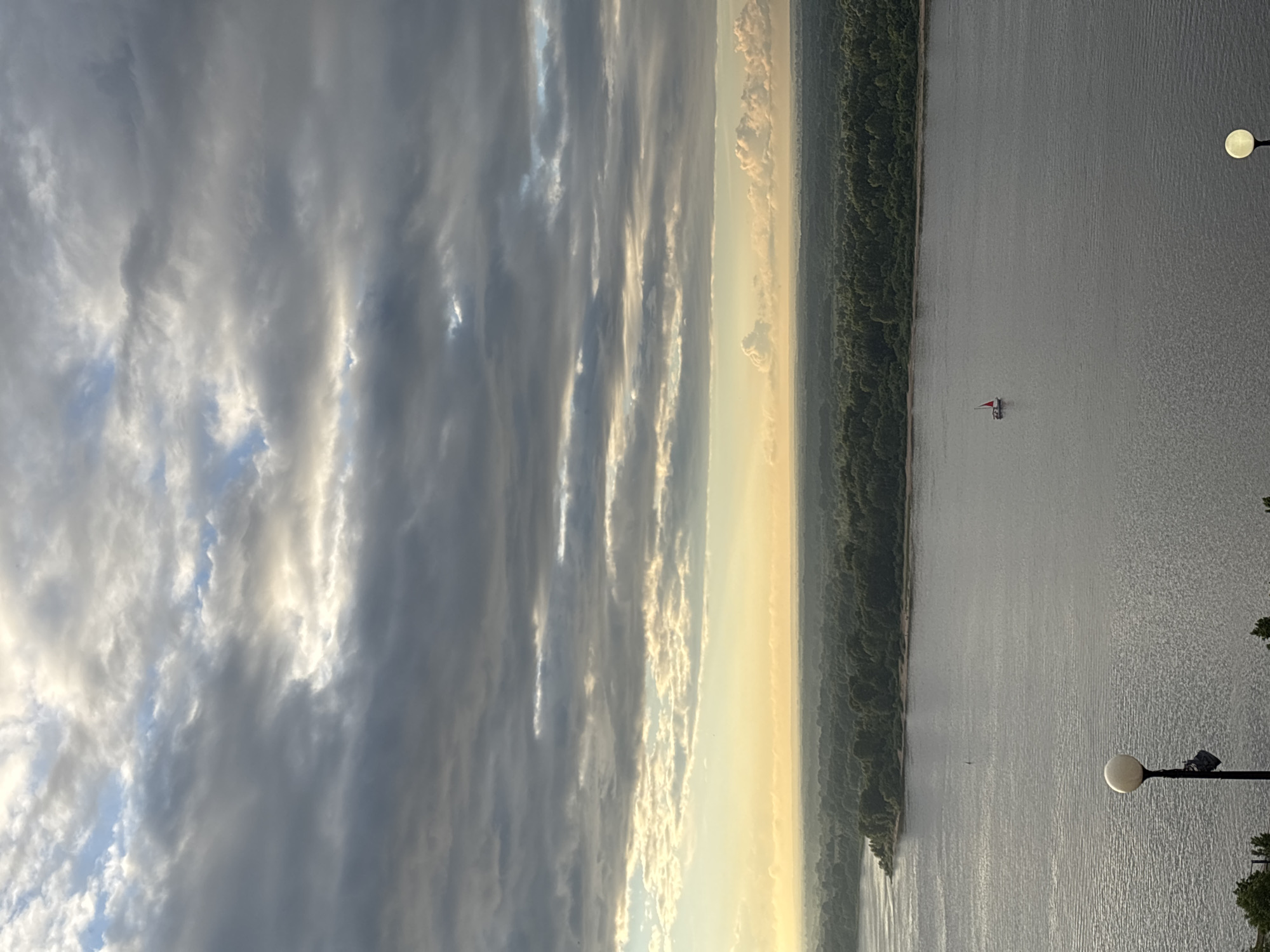 This is a scenic view of the Volga River in Nizhny Novgorod, Russia, at sunrise or sunset. The sky is filled with dramatic, layered clouds, with patches of soft, warm light breaking through, suggesting either the dawn or dusk. Below the sky, a broad, calm river dominates the foreground, its surface reflecting the muted light. On the far bank of the river, a dense, green forest stretches along the shoreline, creating a natural border. In the distance, a small sailboat with a red sail navigates the water. In the foreground, a lamppost with a spherical light fixture is visible on the left side of the image, and another similar fixture is partially visible on the right. The overall atmosphere is serene and picturesque, capturing a moment of natural beauty.