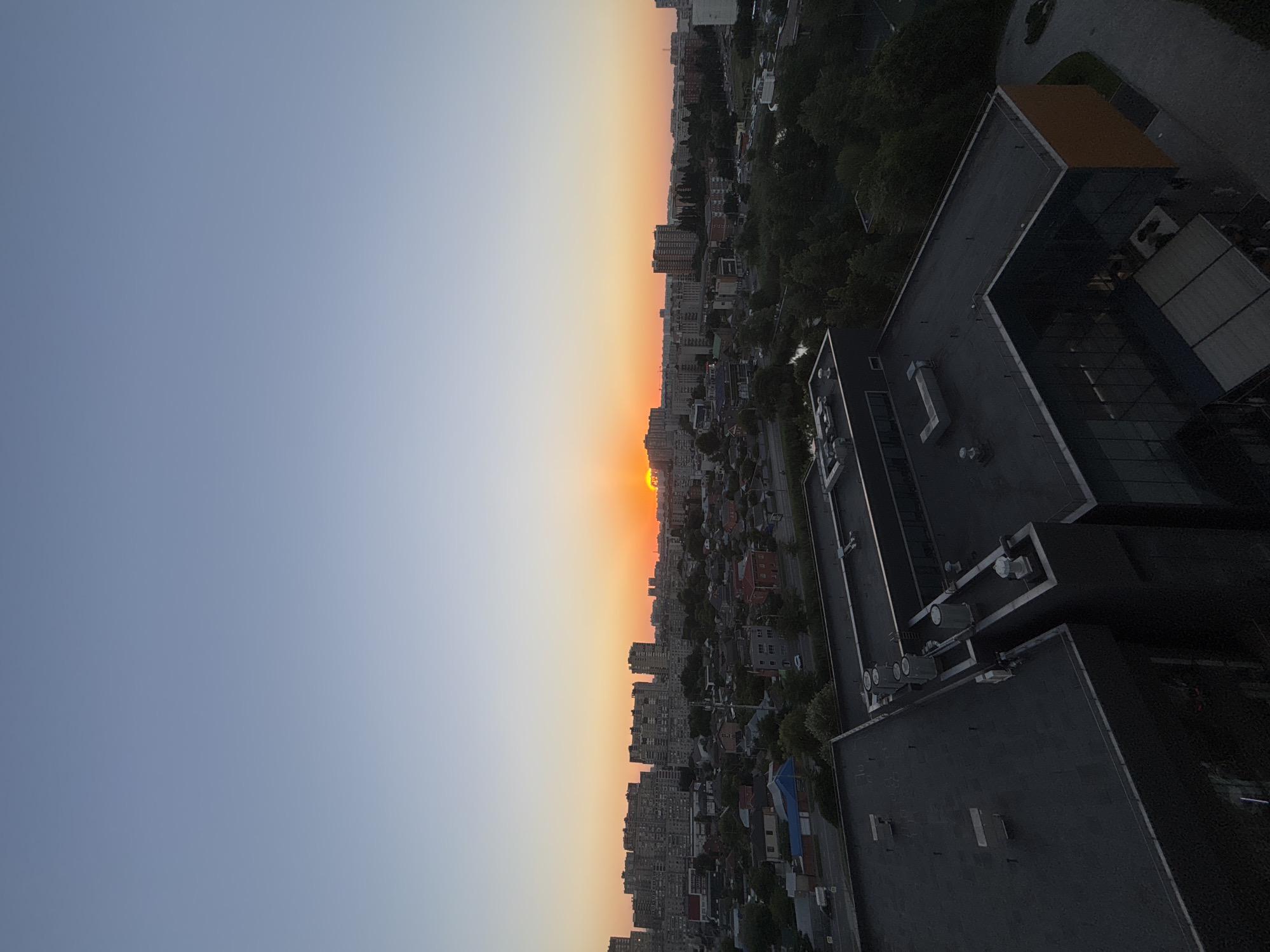 The image captures a cityscape at sunset, viewed from a high vantage point. The sky transitions from a clear, pale blue on the left to a warm gradient of orange and yellow towards the horizon on the right, indicating the sun is setting. Below the sky, a dense urban landscape unfolds with numerous apartment buildings and smaller structures. Trees are interspersed throughout the city, adding touches of green. In the foreground, the dark, flat roofs of modern buildings are visible, featuring various rooftop equipment such as vents and possibly HVAC units. The overall scene depicts a typical urban environment during the golden hour. The time of day is clearly sunset, and the weather appears clear, contributing to the vibrant sky colors. The location is identified as Krasnodar, Russia. No people are clearly discernible in the image, but the presence of buildings and infrastructure suggests human habitation and activity.